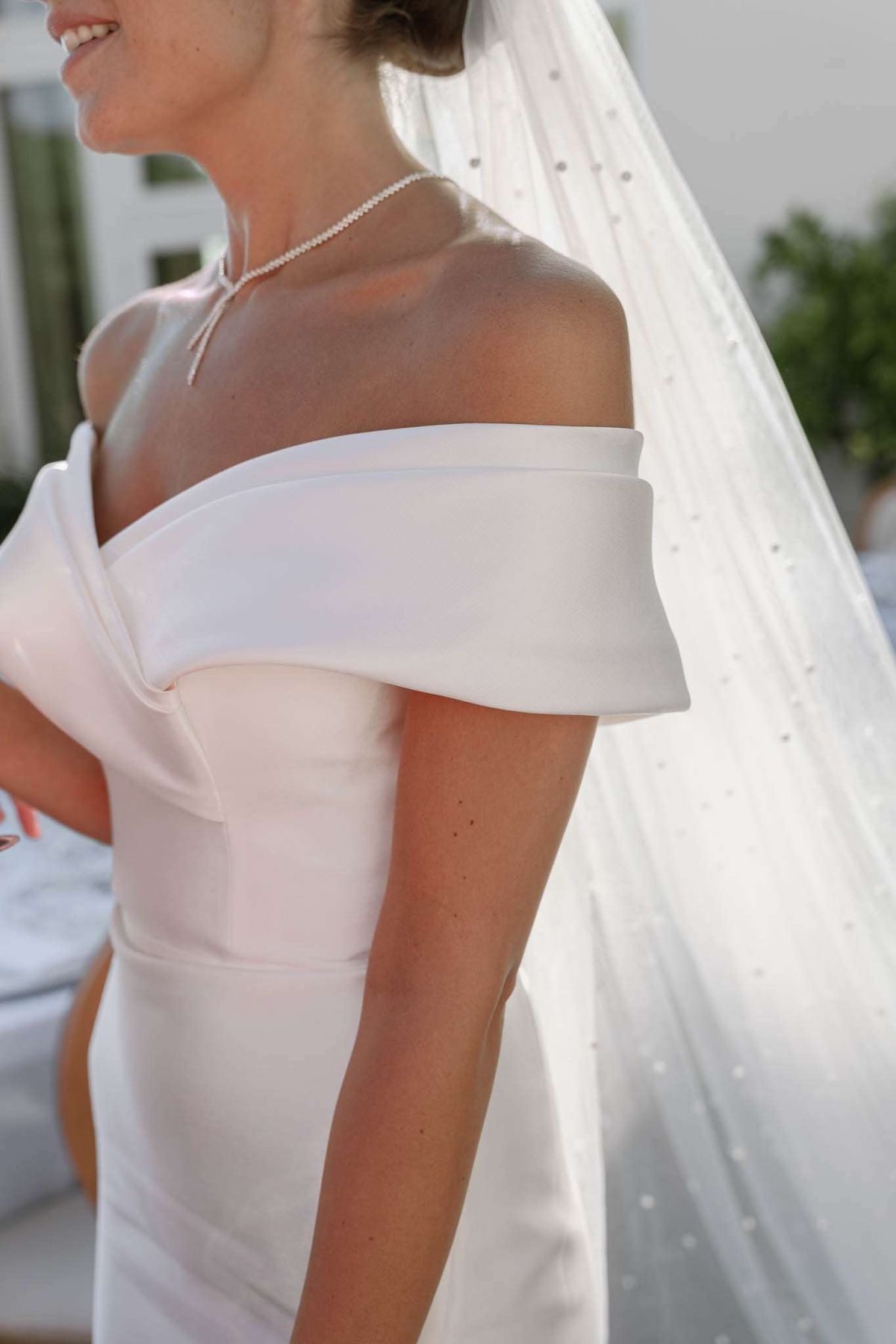 Close-up of bride in off-the-shoulder satin gown with pearl necklace and crystal-dotted veil