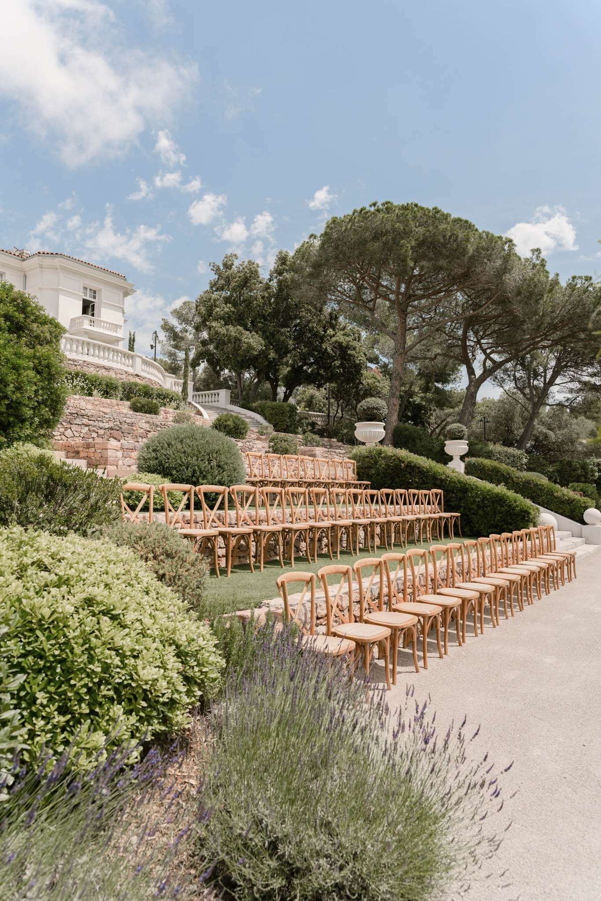 Outdoor ceremony setup with cross-back chairs on terraced grounds of a white Provencal villa with lavender borders