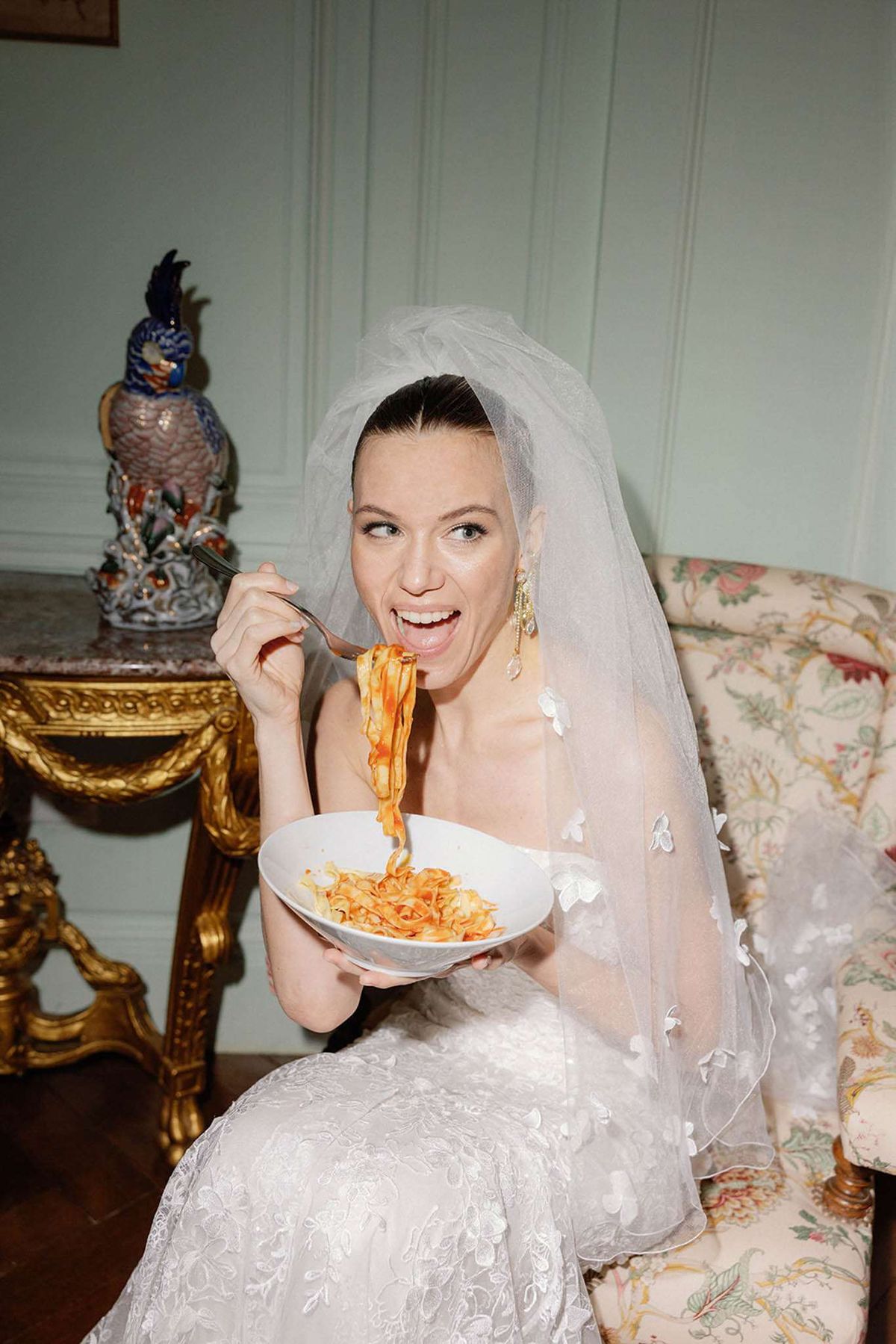 Bride in strapless ivory lace gown laughing while eating pasta on floral sofa inside chateau salon