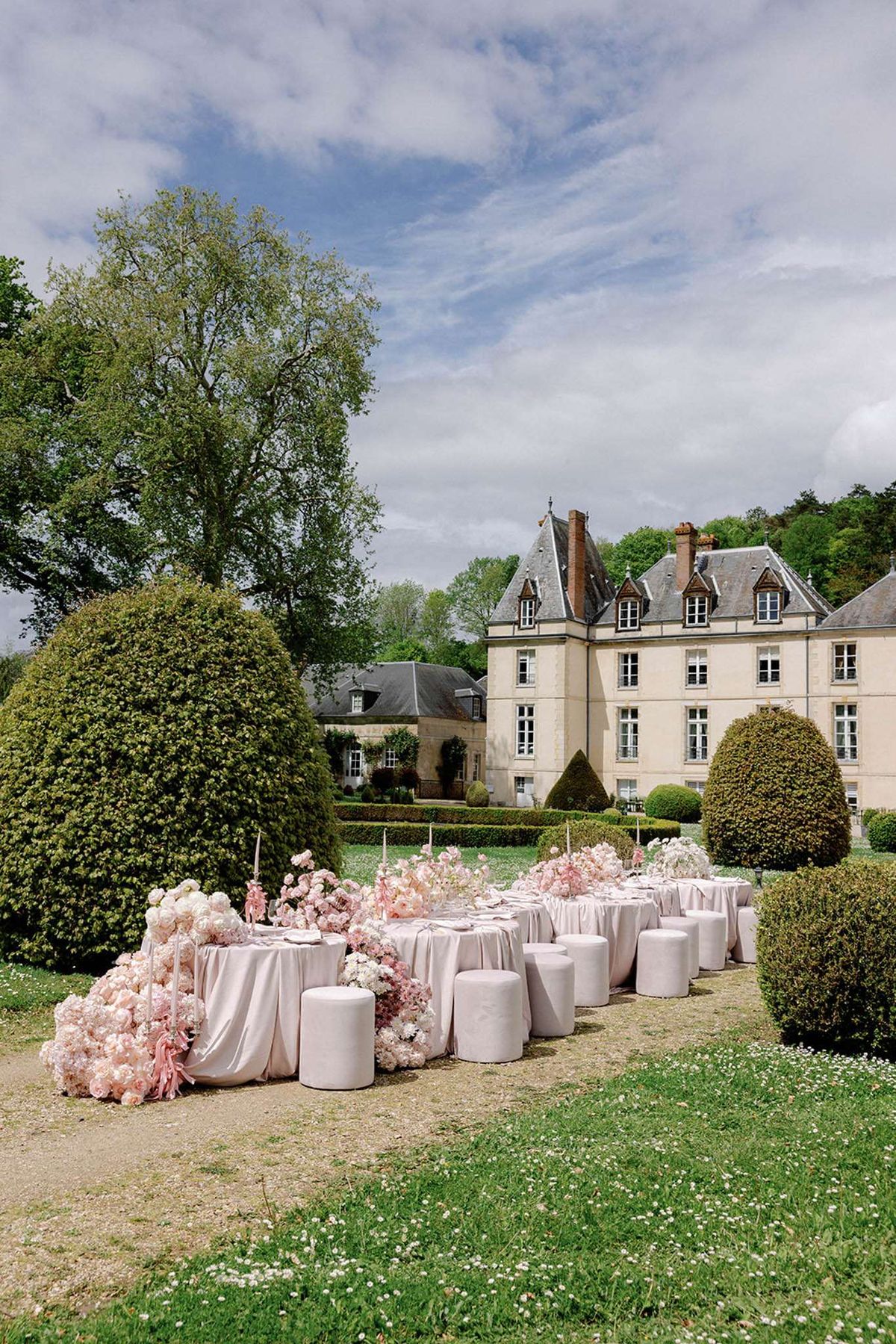 An outdoor wedding reception tablescape is set up in the formal gardens of a French château, with the cream stone manor house and its slate-roofed turrets visible in the background. The long rectangular dining table is dressed in blush pink satin-style linen and flanked by cylindrical upholstered poufs in a matching blush tone, used as seating in place of traditional chairs. The table is heavily decorated with large arrangements of blush pink and ivory flowers — including what appear to be peonies, garden roses, and ranunculus — cascading abundantly along the table's length, with tall tapered pink candles interspersed throughout. The overall decor palette is monochromatic blush pink and ivory, with a modern-romantic styling approach. The image is a wide shot taken from ground level, capturing both the full table setup and the château façade. Potential venue feature image.
