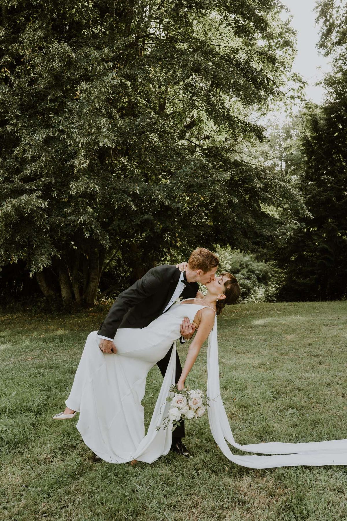 Groom dipping bride for a kiss on garden lawn, bride holding blush rose bouquet with trailing ribbon