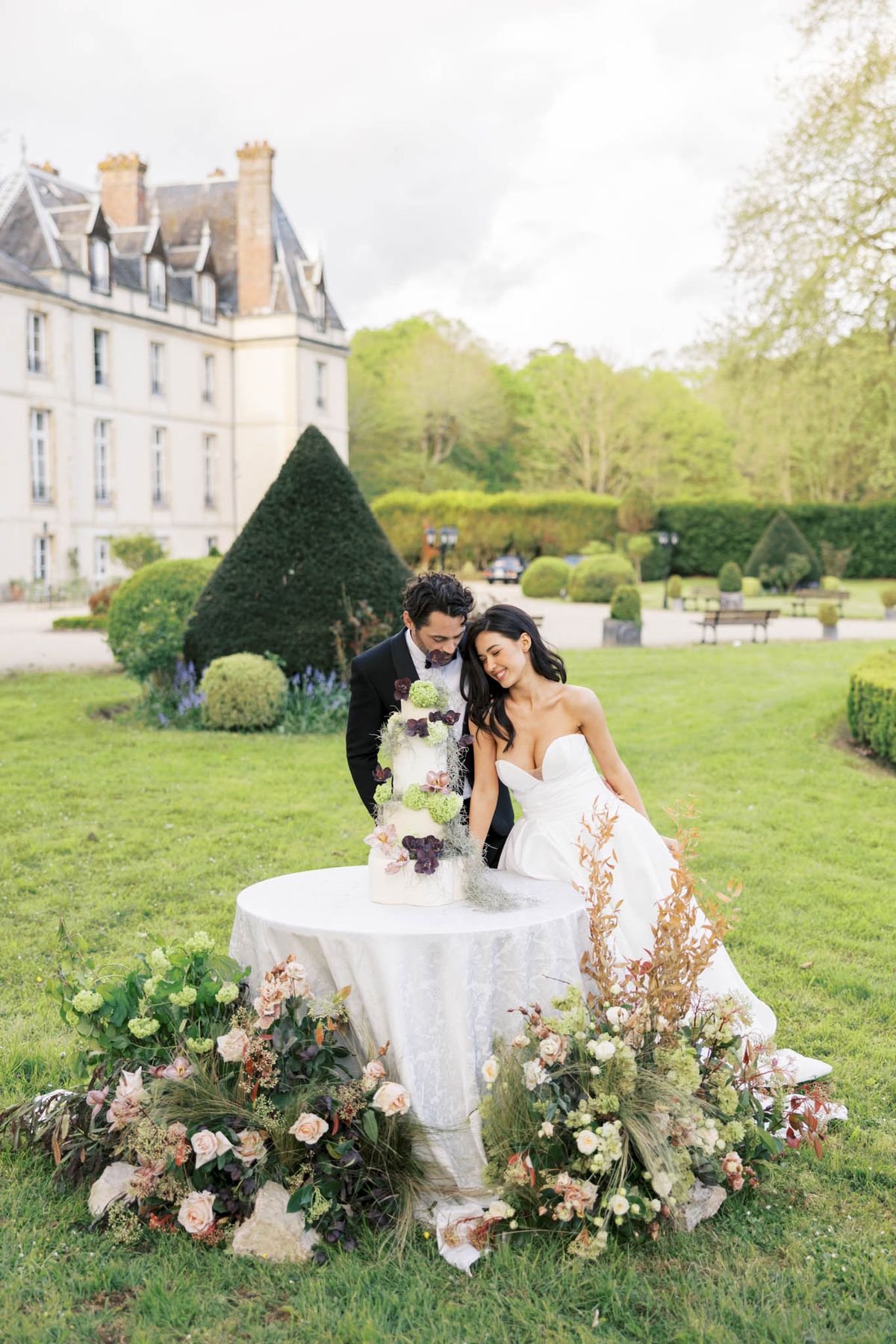 A couple cuts their wedding cake outdoors on the grounds of a French château, with the multi-story cream-stone building with steep slate roofs visible in the background. The bride wears a strapless white ballgown with a structured bodice and full skirt, and the groom is dressed in a black tuxedo with a bow tie; both lean toward each other smiling. The four-tier white cake is decorated with deep burgundy hellebores, chartreuse green viburnum, trailing silver-grey dried grasses, and dark chocolate-toned botanicals. The round cake table is draped in a white textured linen and surrounded at the base by a lush ground-level floral arrangement featuring blush garden roses, white ranunculus, burgundy hellebores, copper-toned foliage, green viburnum, and wild trailing greenery spread across the lawn. The shot is a wide portrait-oriented image taken at ground level, capturing the couple, cake, and château in a single frame.