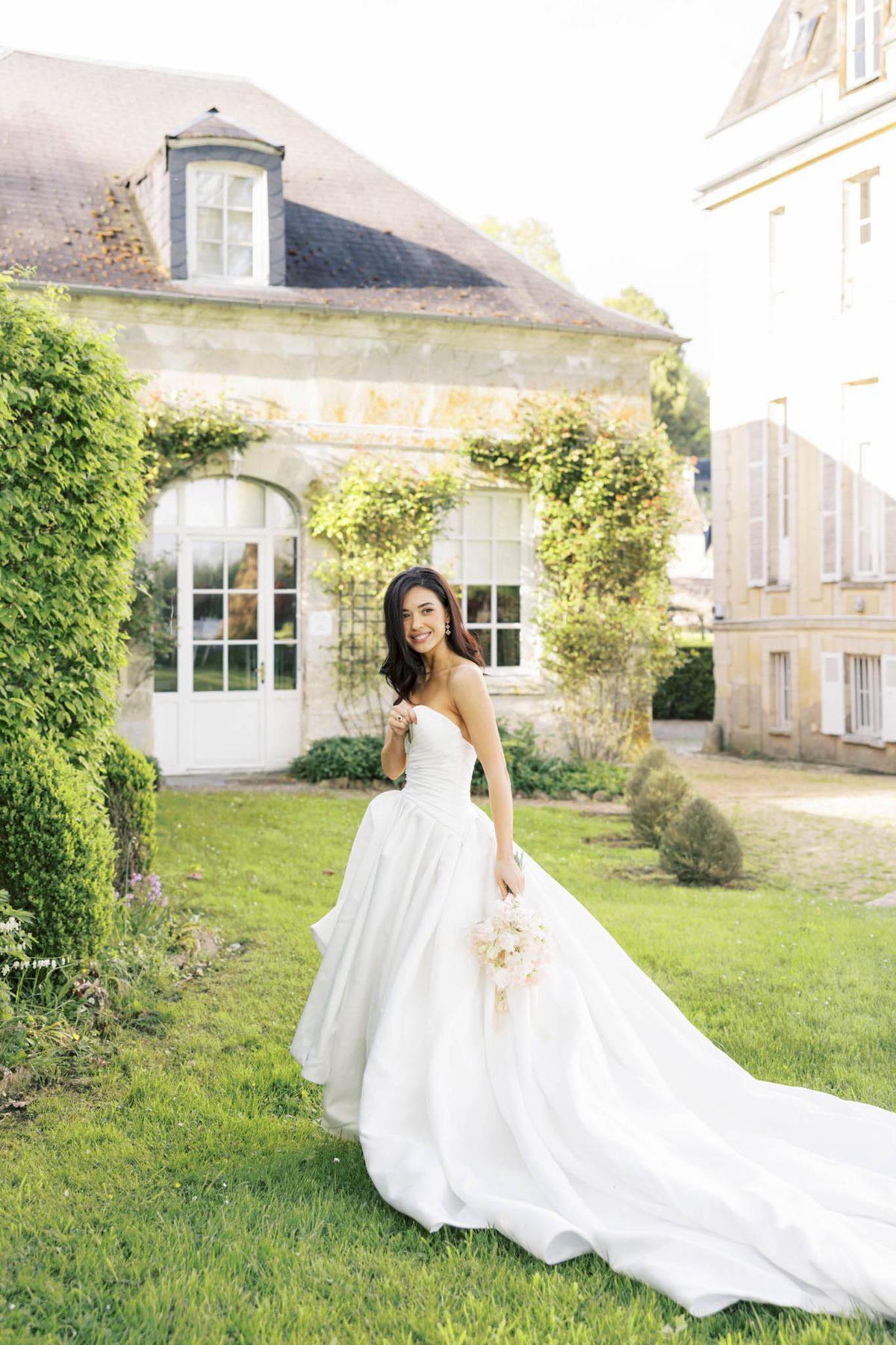 Bride in strapless white ball gown smiling over her shoulder on the lawn of an ivy-covered French chateau