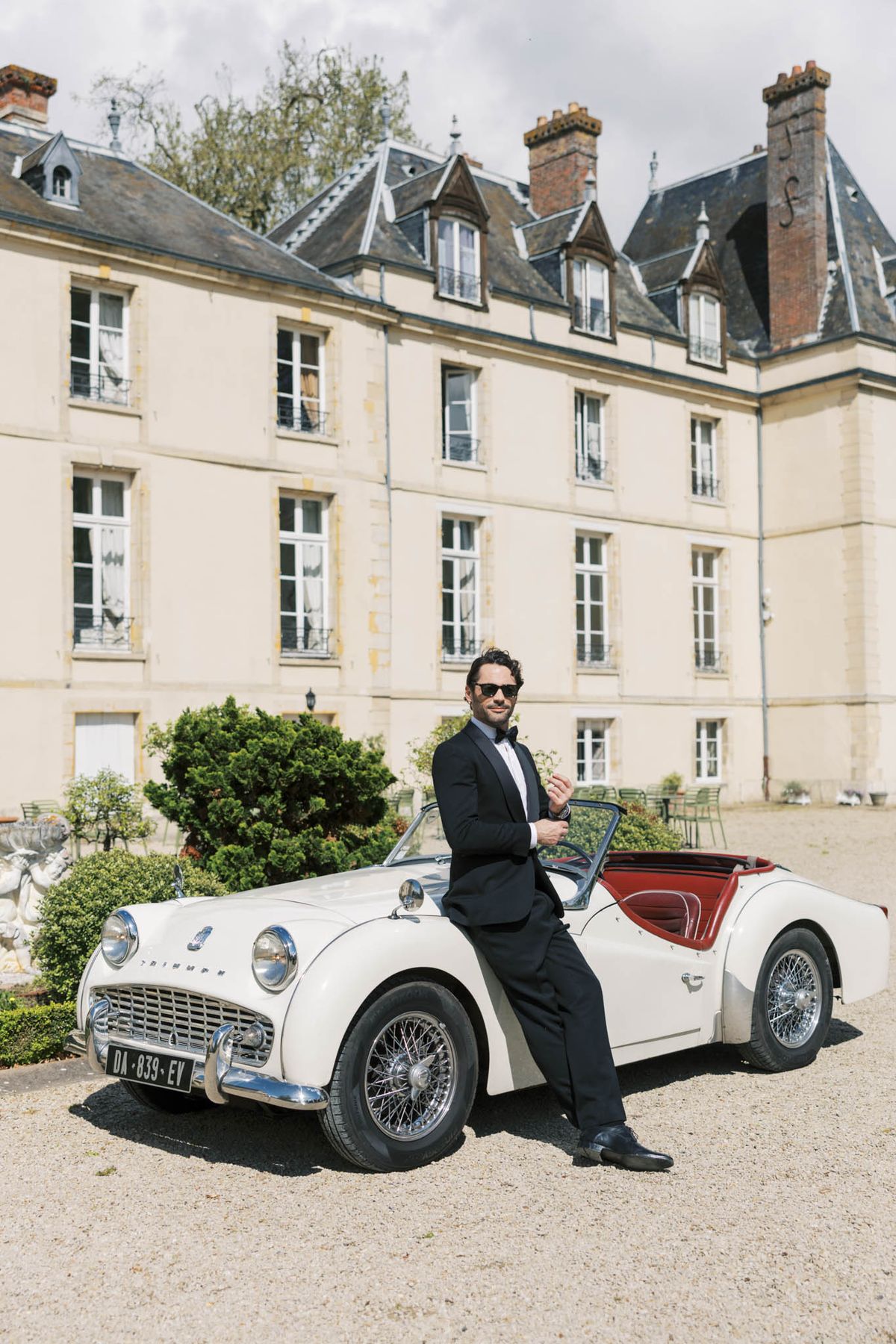 Groom in black tuxedo leaning against vintage cream Triumph convertible in front of French chateau