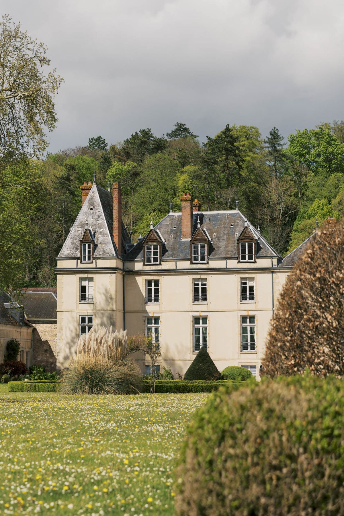 A wide exterior shot of a French château photographed from across the grounds. The building is a three-storey manor house with cream-rendered walls, a slate mansard roof featuring multiple dormer windows with dark wood trim, and several brick chimney stacks. The formal garden in front of the château includes clipped box hedging, a cone-shaped topiary, and ornamental grasses. No people are visible in the frame. Potential venue feature image.