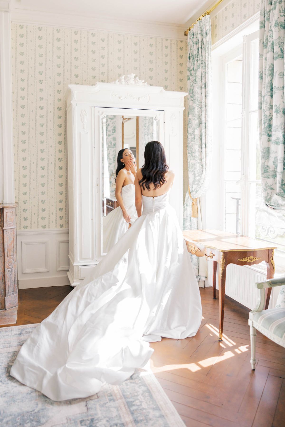 Bride from behind in ivory ballgown with cathedral train examining reflection in armoire mirror in toile room