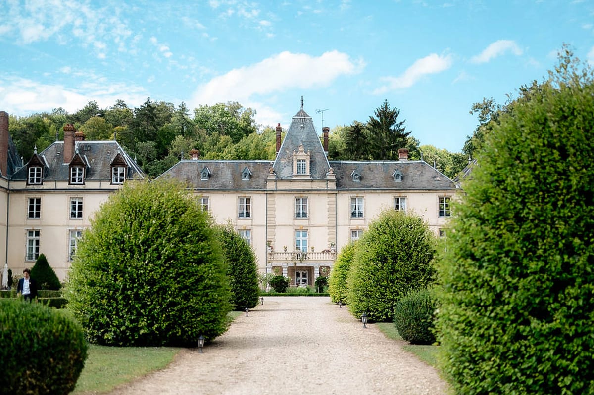 Cream stone chateau with slate mansard roof and central tower approached by lantern-lined gravel path