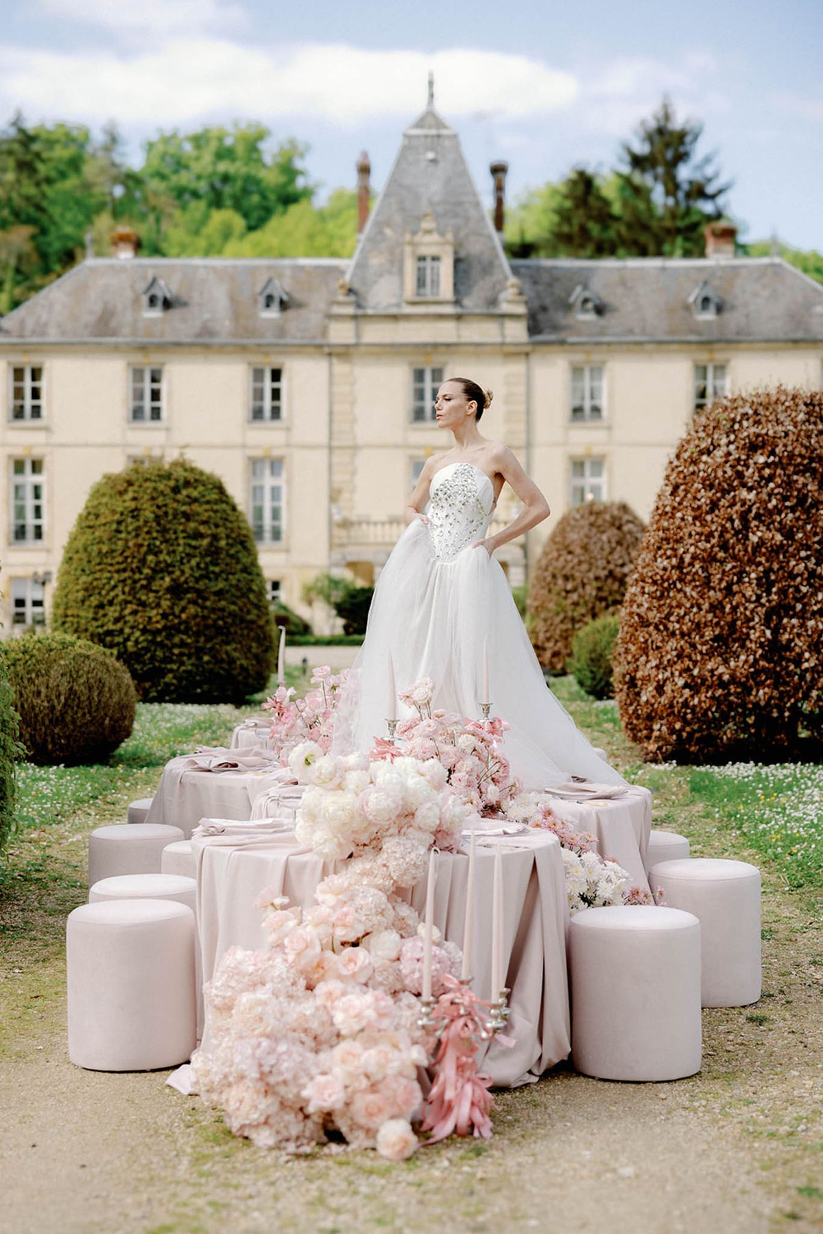 A bride stands behind a styled outdoor reception table set up in front of a classic French château with a steeply pitched slate roof, dormer windows, and manicured topiary. She wears a strapless white ballgown with a heavily crystal-embellished corset bodice and a voluminous skirt, with her hair pulled back in a sleek updo. The long rectangular low table is draped in blush-pink linen and surrounded by cylindrical blush velvet ottomans used as seating; the table runner is an abundant cascade of blush garden roses, ivory peonies, white ranunculus, and pink cherry blossom branches, spilling off both ends onto the ground, with silver taper candelabras and dusty rose ribbon accents interspersed throughout. The shot is a wide full-length portrait taken at ground level, with the château facade centered in the background, placing the bride as the focal point between the table and the building. Potential venue feature image.