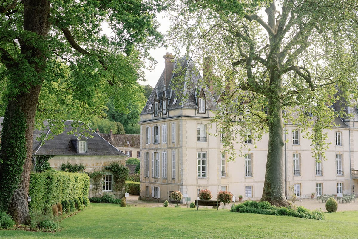 A wide exterior shot of a French château and its surrounding grounds, with no people visible. The main building features cream-colored stone facades, slate mansard roofs with dormer windows, and blue-grey shuttered windows across multiple stories. A prominent corner tower anchors the central section of the property. To the left, a smaller stone outbuilding with a pitched roof and climbing vines is partially visible, framed by a neatly trimmed hedge. The grounds include a gravel courtyard with a wooden bench, potted flowering shrubs in pink and red tones, clipped box balls, and outdoor dining furniture arranged near the château's terrace. The composition is a medium-wide shot taken from the lawn, framed by two large mature trees in the foreground. Potential venue feature image.