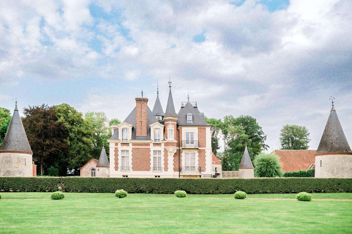 Red brick French chateau with slate turrets, stone detailing, wrought iron balcony, and formal hedgerow garden