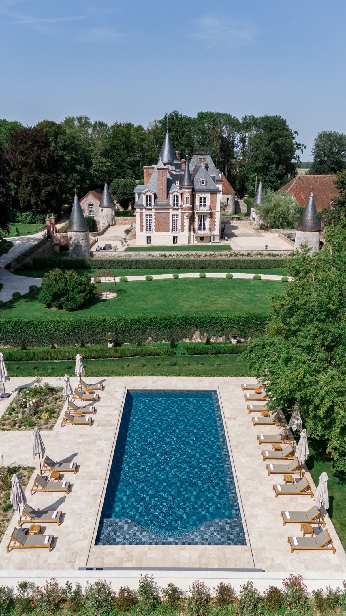 Aerial multi-turreted brick chateau with formal box garden swimming pool limestone terrace and teak loungers