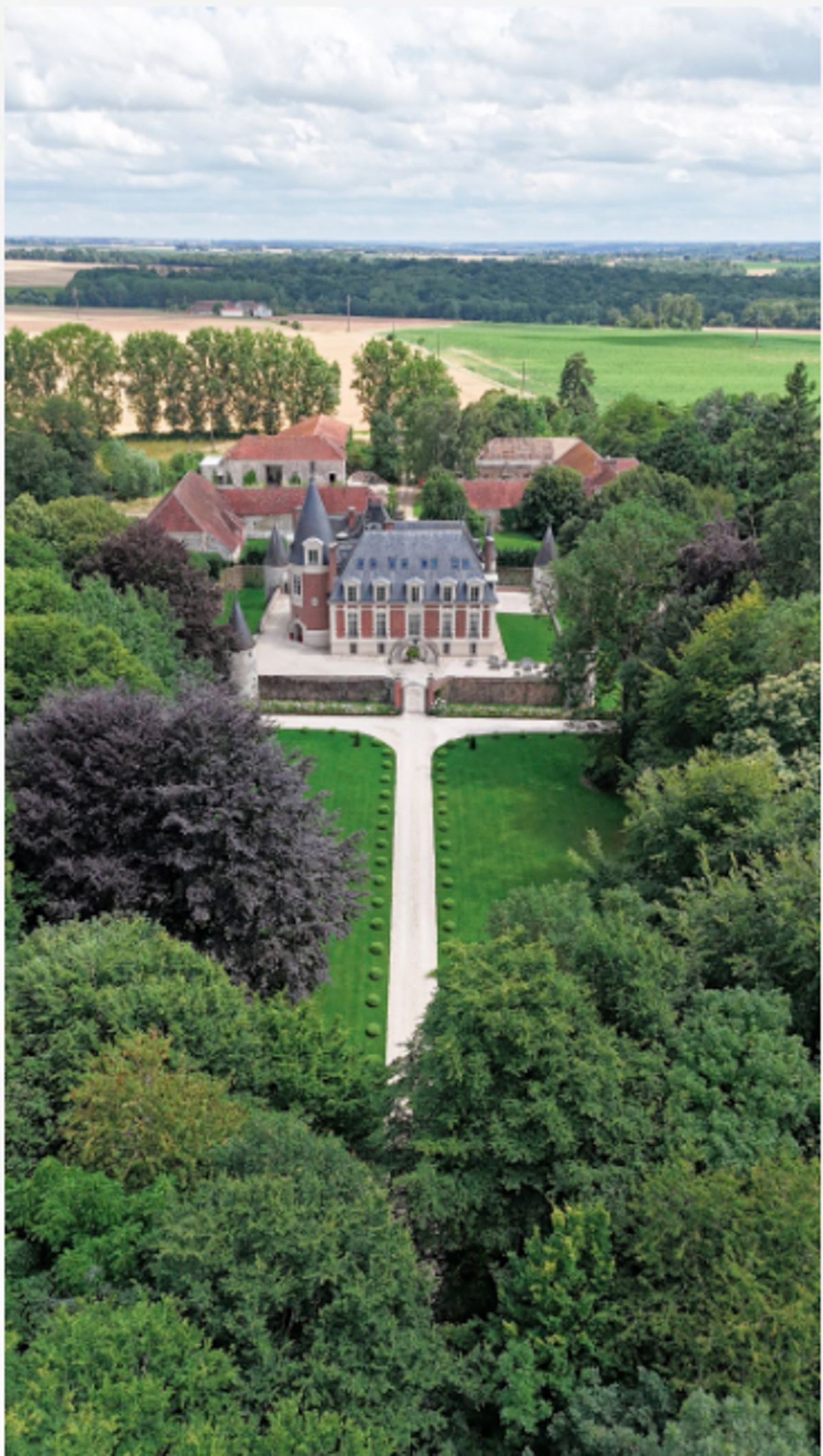 Aerial drone shot of a French château and its surrounding estate, with no wedding party or guests visible. The main building features red brick façades, a slate mansard roof, and conical corner turrets, flanked by outbuildings with terracotta-tiled roofs. A straight gravel driveway leads from the foreground tree line directly to the château's front courtyard, flanked by manicured lawns. The property is set within an extensive wooded park, with open agricultural fields visible in the background. Potential venue feature image.