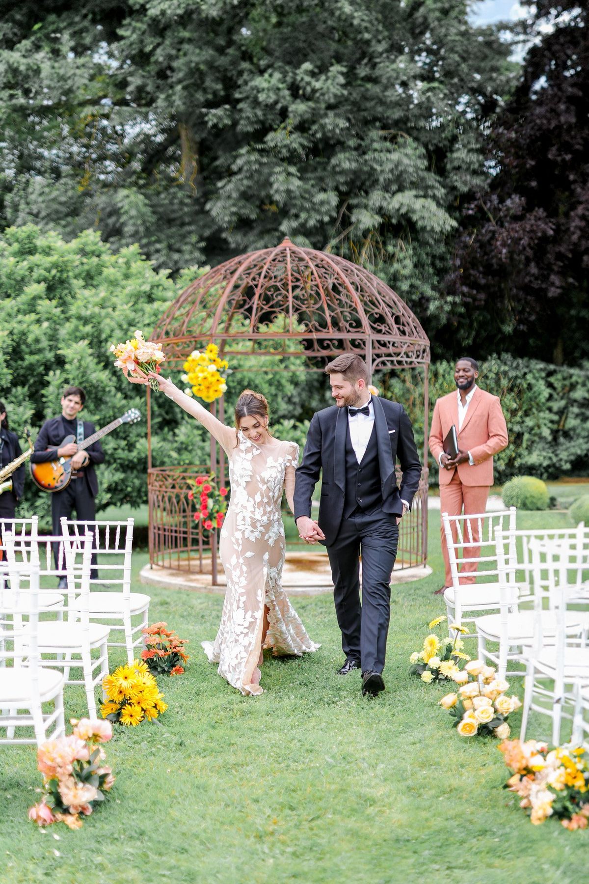 Bride and groom walking back down the aisle after ceremony with raised peach bouquet and guitarist behind