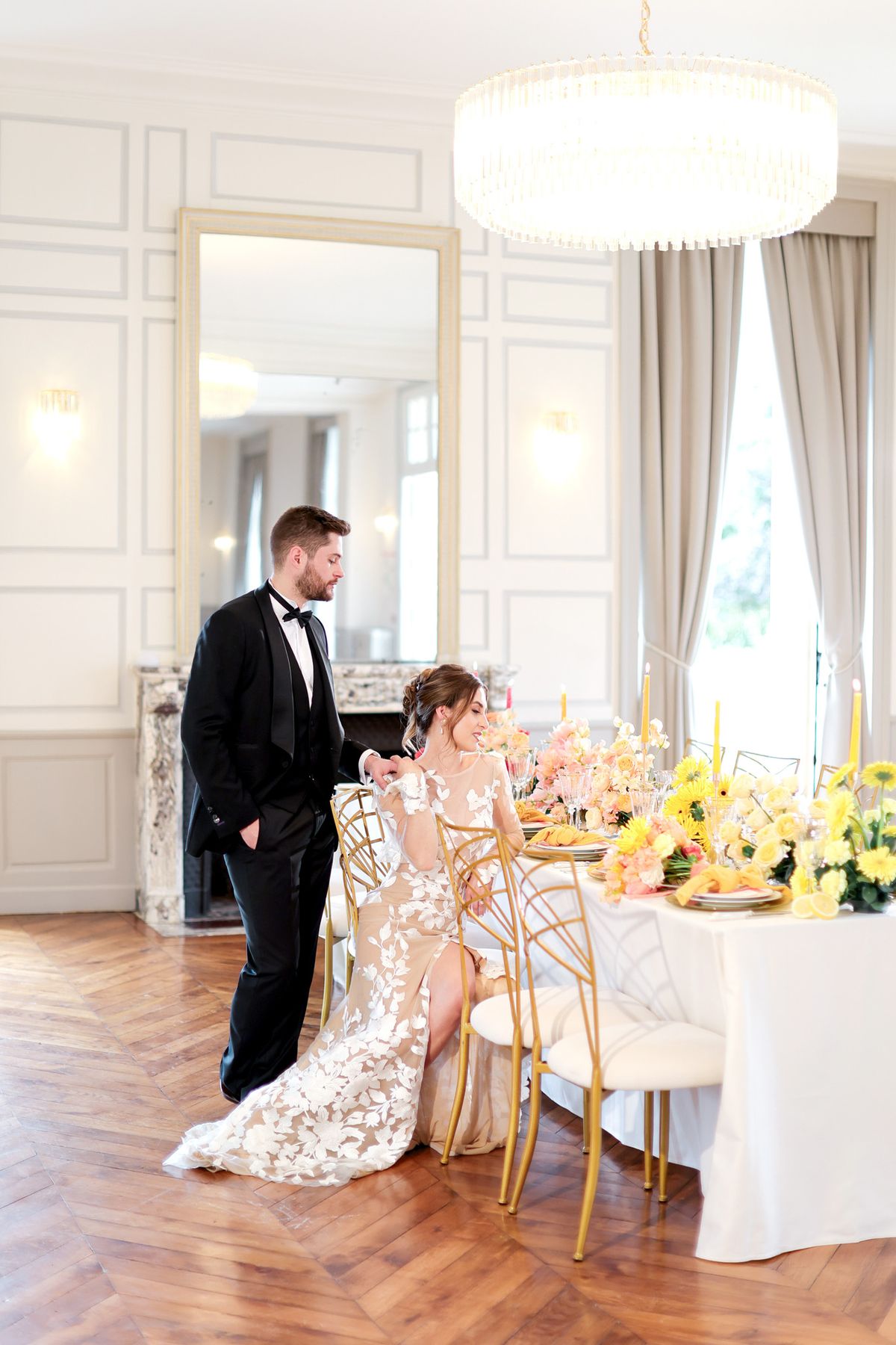A couple poses beside a styled reception table inside a formal French interior room featuring white boiserie wall panelling with gold trim, a marble fireplace, herringbone parquet flooring, and a large floor-length mirror. The groom stands in a black tuxedo with a black bow tie, while the bride is seated in a gold geometric chair wearing a long-sleeved fitted gown with a nude illusion base and white three-dimensional floral lace appliqué. The tablescape features a white linen-covered table dressed with yellow taper candles, yellow chrysanthemums, coral and peach blooms, gold-rimmed place settings, and crystal glassware, styled in a modern classic palette of yellow, peach, and gold. A large cylindrical crystal chandelier hangs overhead, and the overall styling is modern classic with a bold warm-toned floral palette. Wide portrait shot capturing the couple and full table setting.