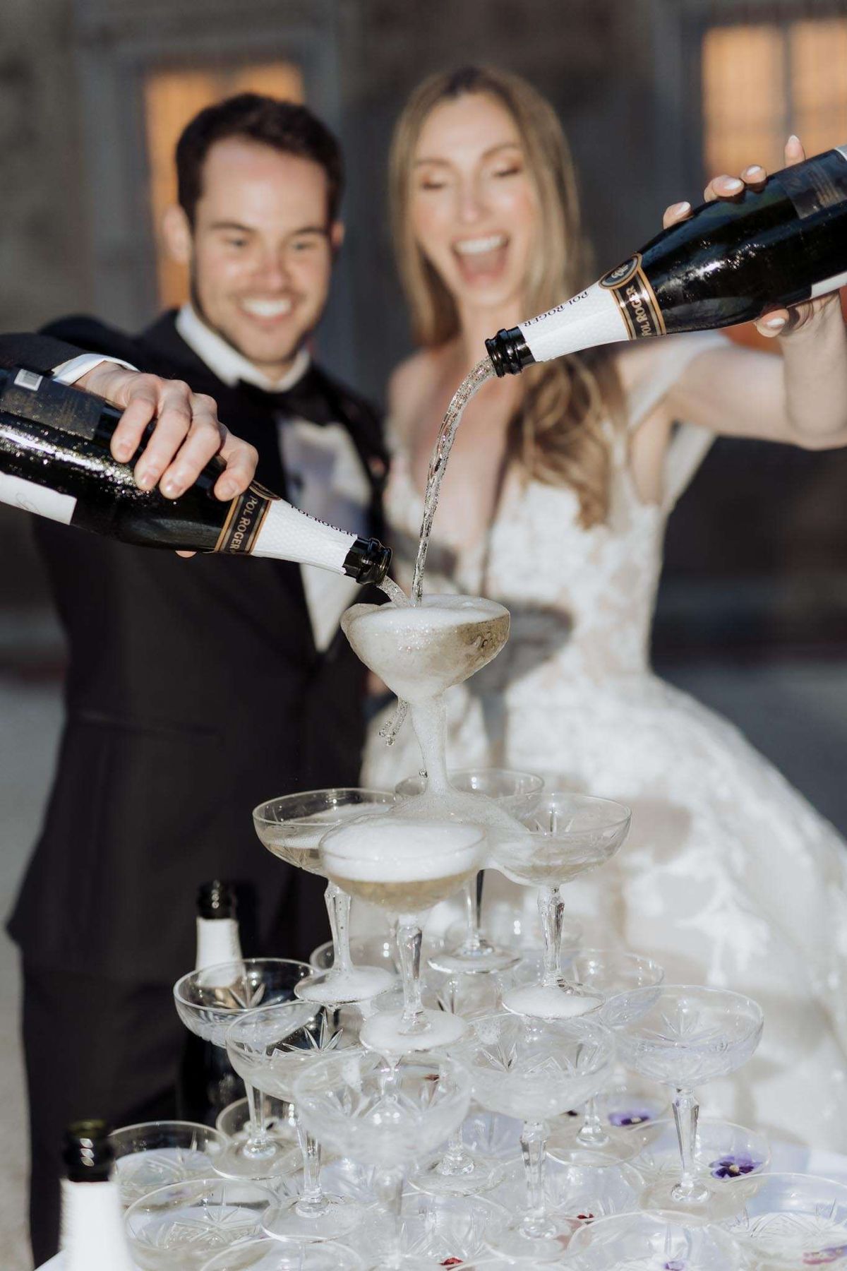 Crystal coupe champagne tower overflowing as couple pours Pol Roger, purple pansies decorating base tier