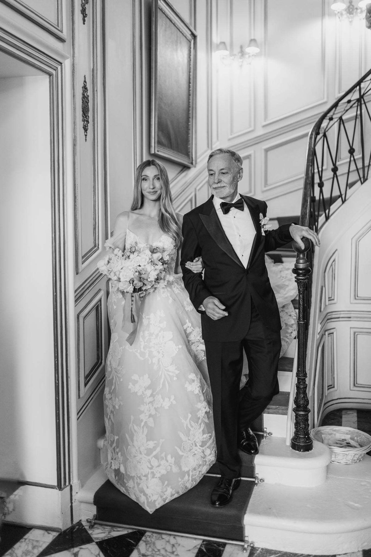 A black-and-white portrait of a bride and an older man, likely her father, preparing to walk down a grand interior staircase at what appears to be a French château or manor house. The bride wears a full ball gown with intricate floral lace appliqué and an off-the-shoulder style, carrying a full, loosely arranged bouquet of roses and soft blooms. The man is dressed in a black tuxedo with a bow tie and a boutonnière on his lapel. The interior features ornate white boiserie wall paneling, framed paintings, a black-and-white checkered marble floor, and a curved wrought-iron staircase railing with a carved wooden newel post, lit by a chandelier in the background. The image is a medium-wide portrait shot with soft contrast and bright highlights on the white tones of the dress and walls.