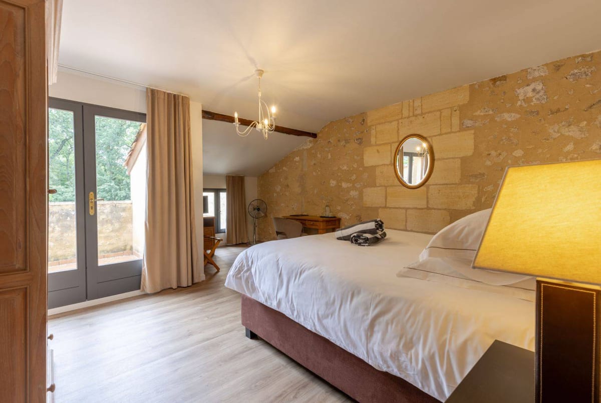 Interior shot of a guest bedroom, likely within a French château or domaine available for wedding accommodation. The room features an exposed golden limestone block wall, a double bed with white linen and a mauve/dusty rose base, light wood flooring, and a white wrought-iron chandelier with candle-style bulbs. A gold-framed oval mirror hangs on the stone wall above the bed, and a warm amber table lamp sits on a dark bedside table. The room includes sand-colored linen curtains, a wooden writing desk and chair, and grey French doors opening to a small terrace. A wooden ceiling beam is visible, giving the space a classic French country aesthetic. Wide-angle interior shot. Potential venue feature image.