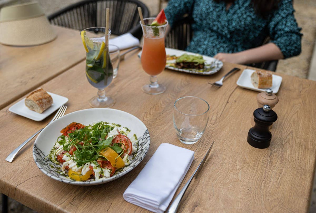 This image does not appear to be a wedding photograph. It shows an outdoor restaurant or café dining scene with a wooden table set for two guests. The foreground features a patterned ceramic bowl with a heirloom tomato and burrata salad topped with fresh arugula, alongside a white linen napkin, cutlery, an empty water glass, and a black pepper mill. Two cocktails are visible — a mint and lemon water-based drink and a pink watermelon-garnished drink in a hurricane glass — along with bread plates at each setting. One person, partially visible, wearing a teal floral blouse, is seated and eating a dish that appears to be a layered sandwich or flatbread. A straw hat rests on a chair in the background. This image does not contain wedding-related content and is not suitable for a wedding content library.