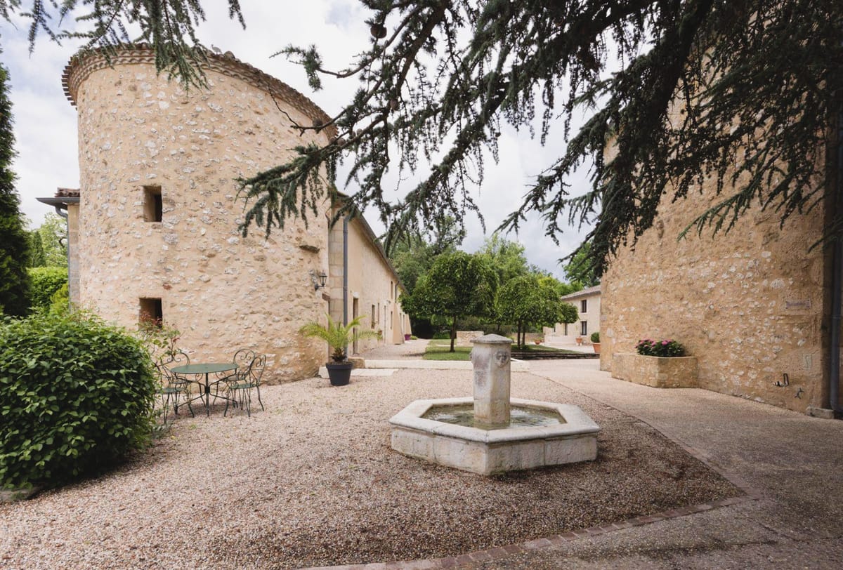 Stone courtyard with round tower, hexagonal fountain, bistro table, and citrus tree row at French estate