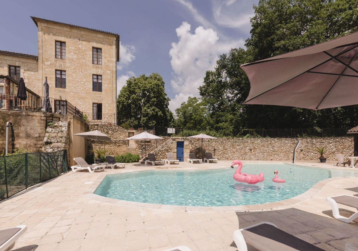 An outdoor pool area at what appears to be a French stone manor or château venue, photographed in bright daytime light with no people present. The kidney-shaped pool features two pink inflatable flamingos floating on the water's surface. The pool terrace is paved with light beige stone slabs and furnished with white and dark grey sun loungers and multiple taupe patio umbrellas. The pool is enclosed by a rustic stone wall with a blue wooden door, and the multi-storey honey-coloured stone manor building is visible to the left. Wide establishing shot. Potential venue feature image.