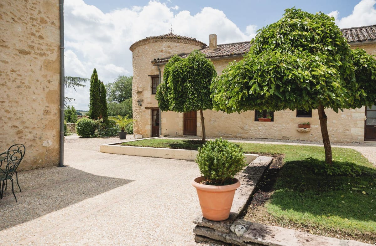A wide exterior shot of a French countryside property featuring a round stone tower with a conical tiled roof and wooden arched doorway, connected to a low stone wing with shuttered windows. The courtyard is gravel-surfaced with a curved planted border containing two topiary-trimmed lollipop trees and a terracotta pot with a clipped box ball. A wrought iron chair is visible on the left edge. The architecture is classic French rural style in warm honey-toned limestone. No people or wedding-specific elements are visible in this image. Potential venue feature image.