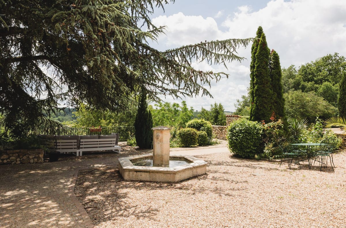 Stone fountain on gravel terrace with bistro chairs, cypress trees, and valley view at French country estate