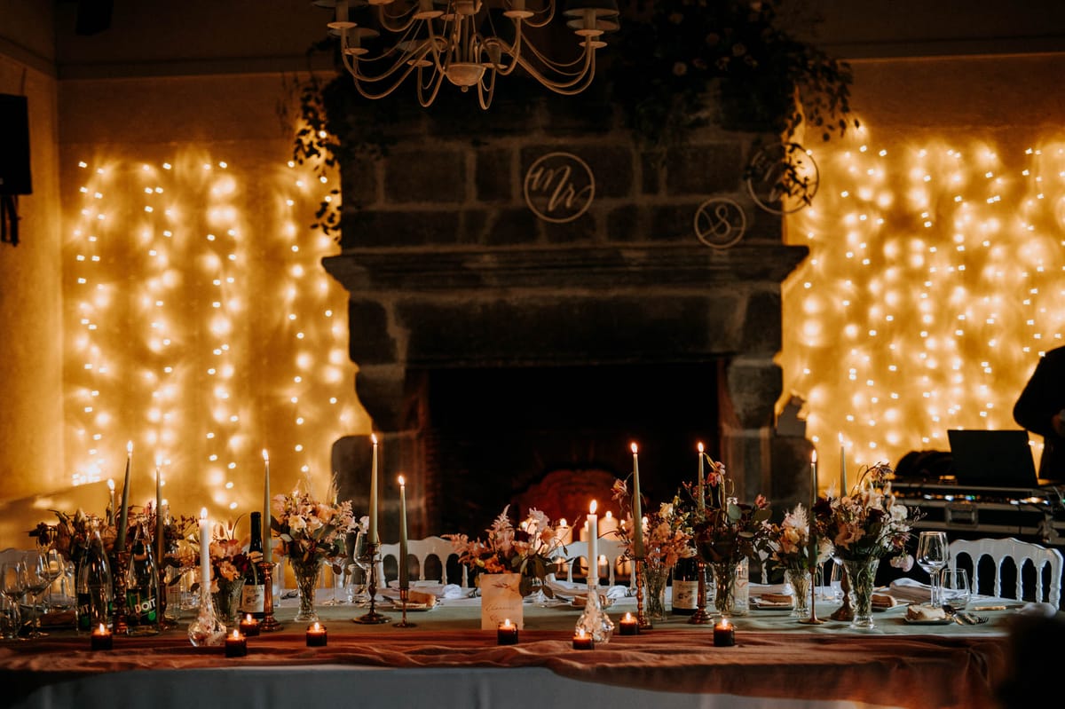Long reception table with terracotta runner, floral centerpieces, sage candles, and fairy-lit stone fireplace backdrop