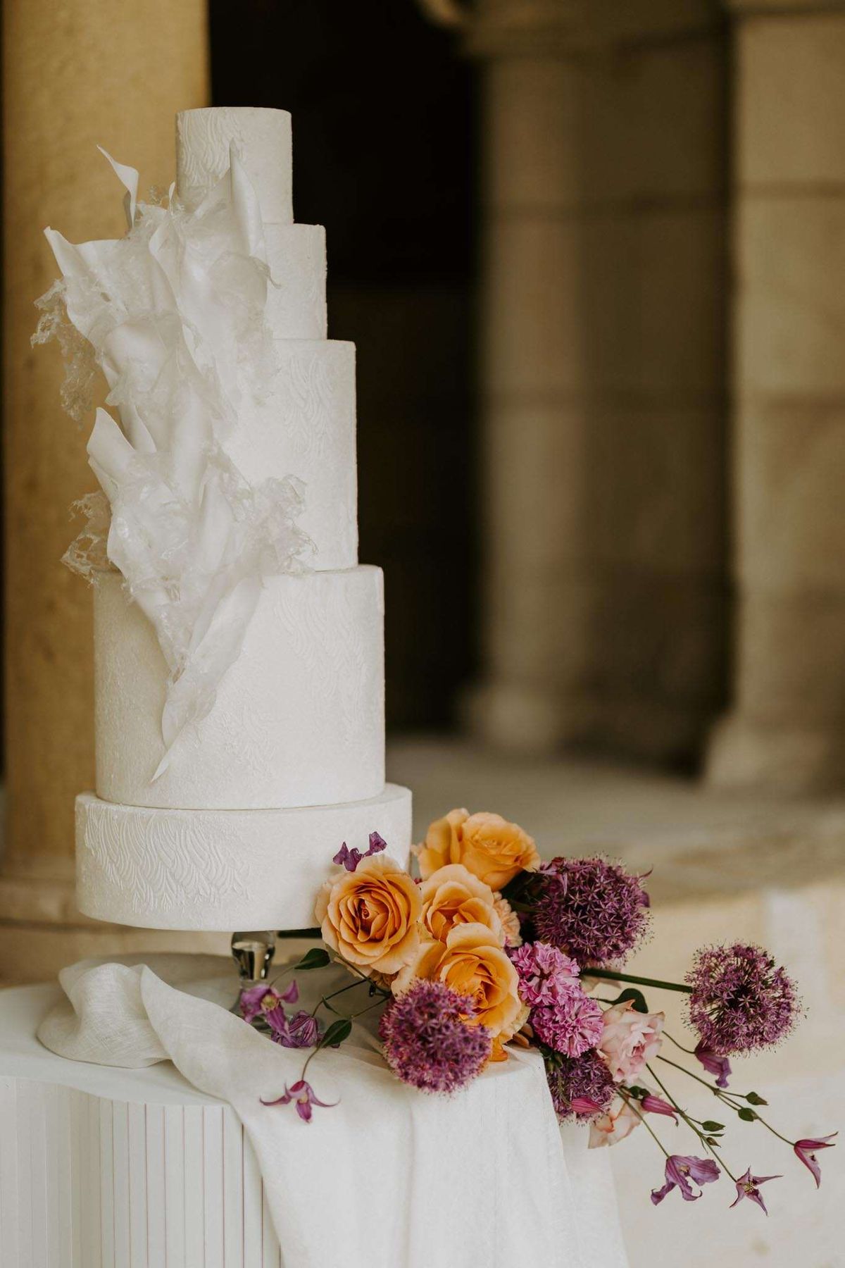 Close-up detail shot of a tall five-tier white wedding cake displayed on a white linen-covered pedestal table, set against the stone columns of what appears to be a classical French château or manor exterior. The cake features an all-white textured finish with an architectural sugar work design — large, irregular white sugar or wafer paper shards cascading down one side alongside what appears to be lace-like sugar detailing. At the base of the cake, a loose arrangement of amber/apricot garden roses, purple allium globes, hot pink carnations, blush roses, and trailing purple clematis blooms is casually placed on the table alongside a draped white silk ribbon. The overall styling is modern and sculptural with a bold floral color palette contrasting against the all-white cake.
