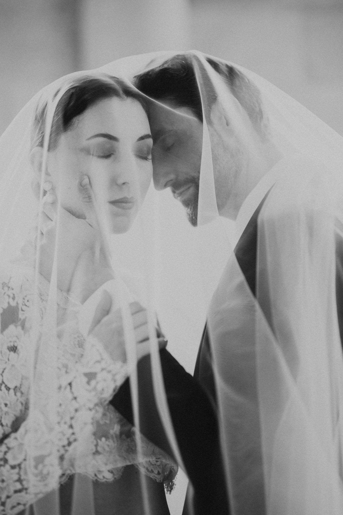 Bride and groom touch foreheads under draped veil with lace sleeve detail visible in soft B&W close-up