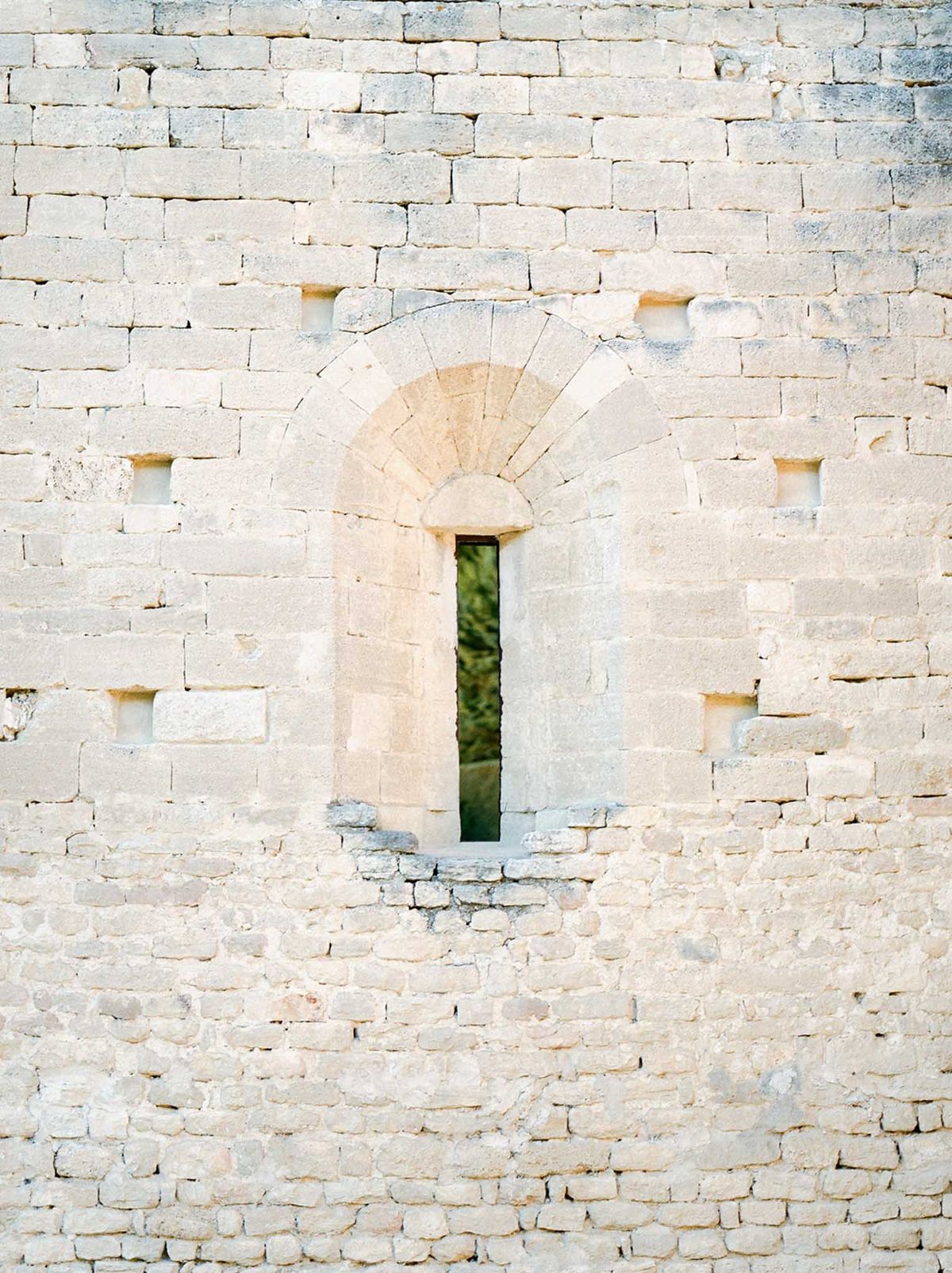 Narrow Romanesque arched window slit in aged limestone wall with green foliage visible through opening