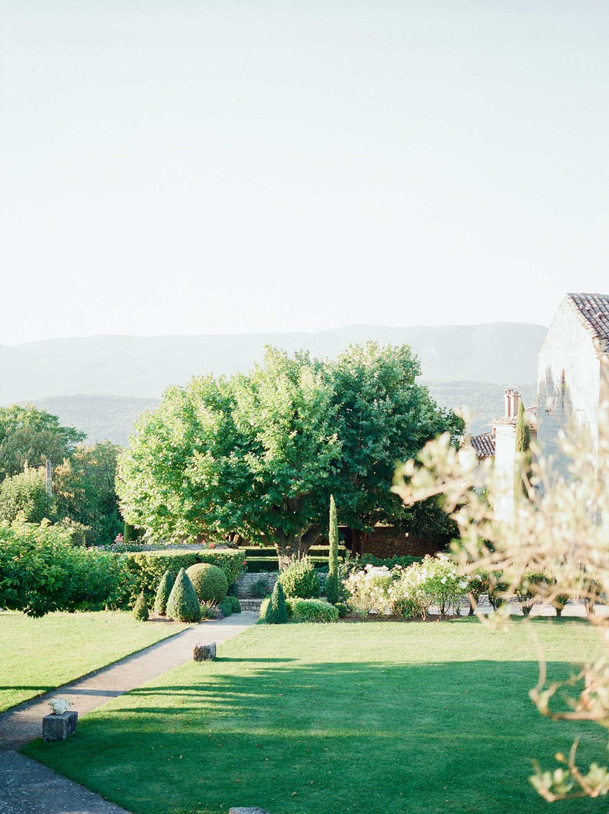 A wide exterior shot of a French estate venue showcasing formal garden grounds with a well-maintained lawn in the foreground, a stone pathway leading toward a structured garden bed featuring clipped topiary cones and rounded box hedges, and a partial view of a stone building with a terracotta tile roof on the right side of the frame. No people are visible in the image. The composition emphasizes the grounds and architecture, with a hazy mountain range visible in the background and tall cypress trees adding vertical structure near the building. The overall styling of the gardens reflects a classic French formal garden aesthetic. Potential venue feature image.