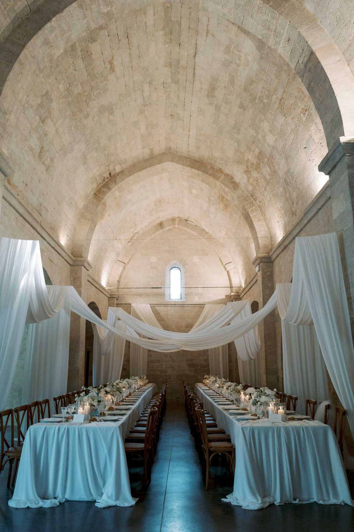 Two parallel tables with sheer canopy drapes in Romanesque vaulted abbey nave with white rose centerpieces