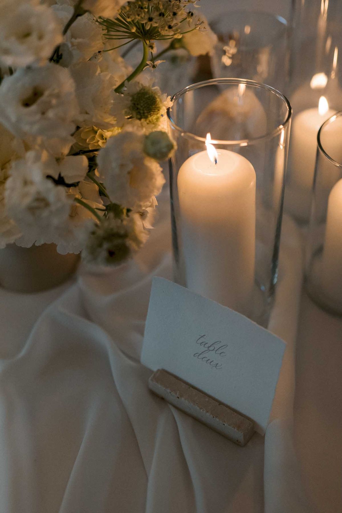 Calligraphy table number card beside white ranunculus arrangement and pillar candles in hurricanes