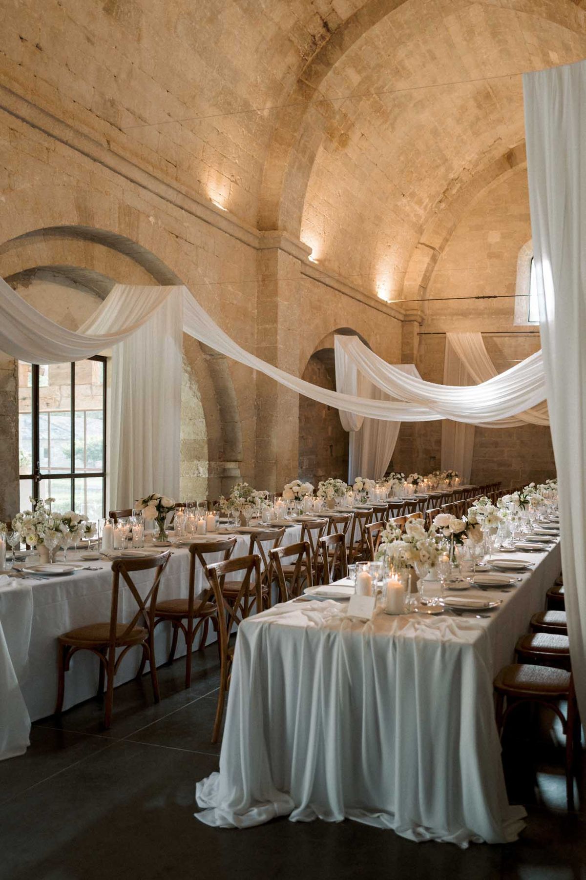 A wedding reception setup inside a vaulted stone hall, likely a historic abbey or monastery, featuring two long parallel banquet tables dressed in floor-length white linen tablecloths. The tables are set with white ceramic plates, crystal glassware, and lit pillar and votive candles, and decorated with low centerpieces of white roses, white hydrangeas, and small white blooms in clear glass vessels. Walnut-toned cross-back wooden chairs line both sides of each table. White sheer fabric is draped in sweeping swags across the ceiling, looping between tall white curtain panels at the corners, creating a soft canopy effect against the warm amber-lit stone vaulted arches above. The overall palette is white, ivory, and warm wood tones, with a classic, refined aesthetic. Wide shot taken from one end of the room looking down the length of the tables. Potential venue feature image.