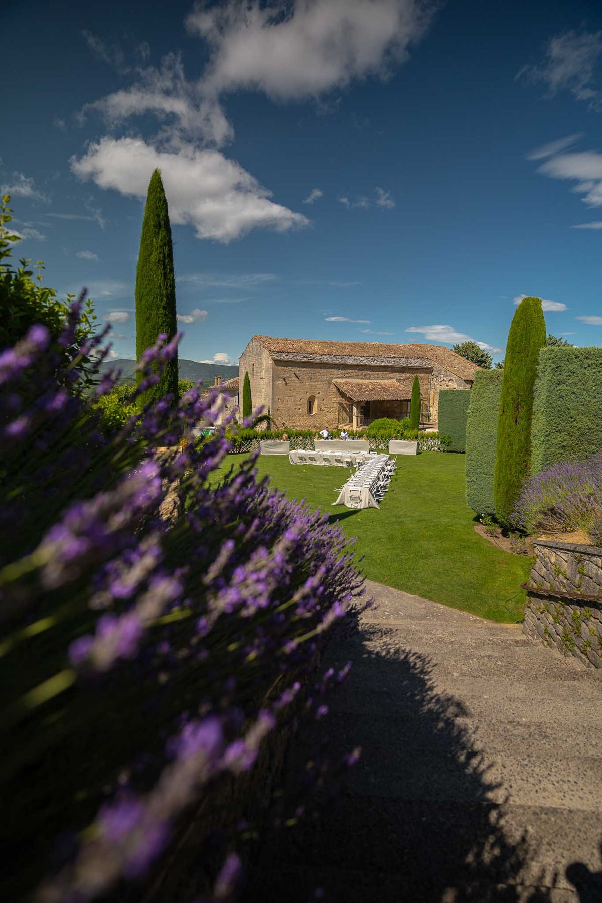 An outdoor ceremony setup is being prepared on a manicured lawn in front of a large stone Provençal farmhouse or mas with a terracotta tile roof. White folding chairs are arranged in two sections with a central aisle, and white-draped tables are positioned at the front near the building. The wide shot is taken from an elevated angle looking down into the garden, with blooming purple lavender plants in the foreground out of focus. Tall cypress trees frame the scene on both sides, and one or two staff members can be seen arranging the setup near the altar area. The overall decor palette is white against the green lawn, with the lavender borders adding a distinctly Provençal character to the setting. Potential venue feature image.