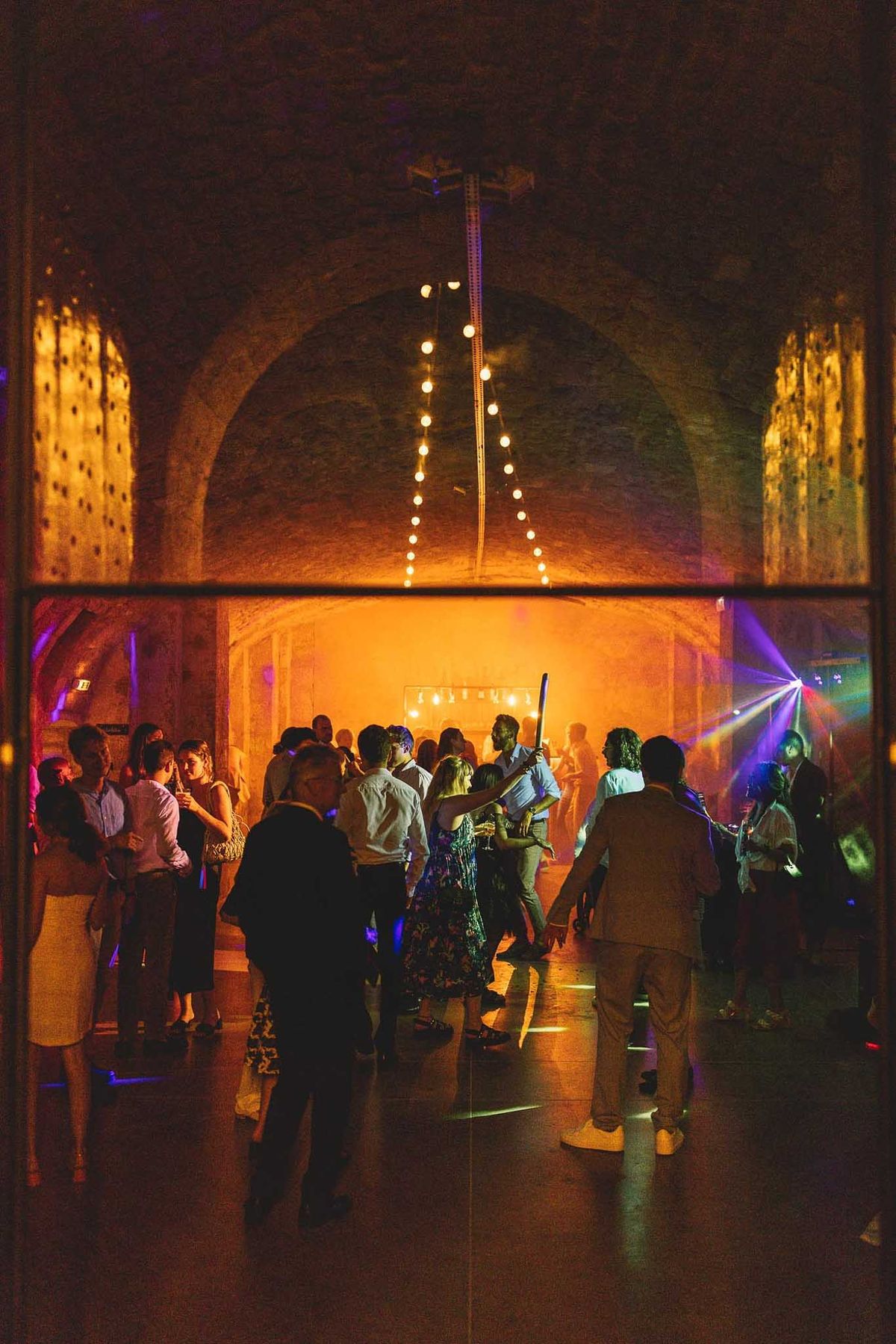 Dance floor in stone-vaulted venue with DJ colored uplighting fog machine fairy lights and thirty dancing guests