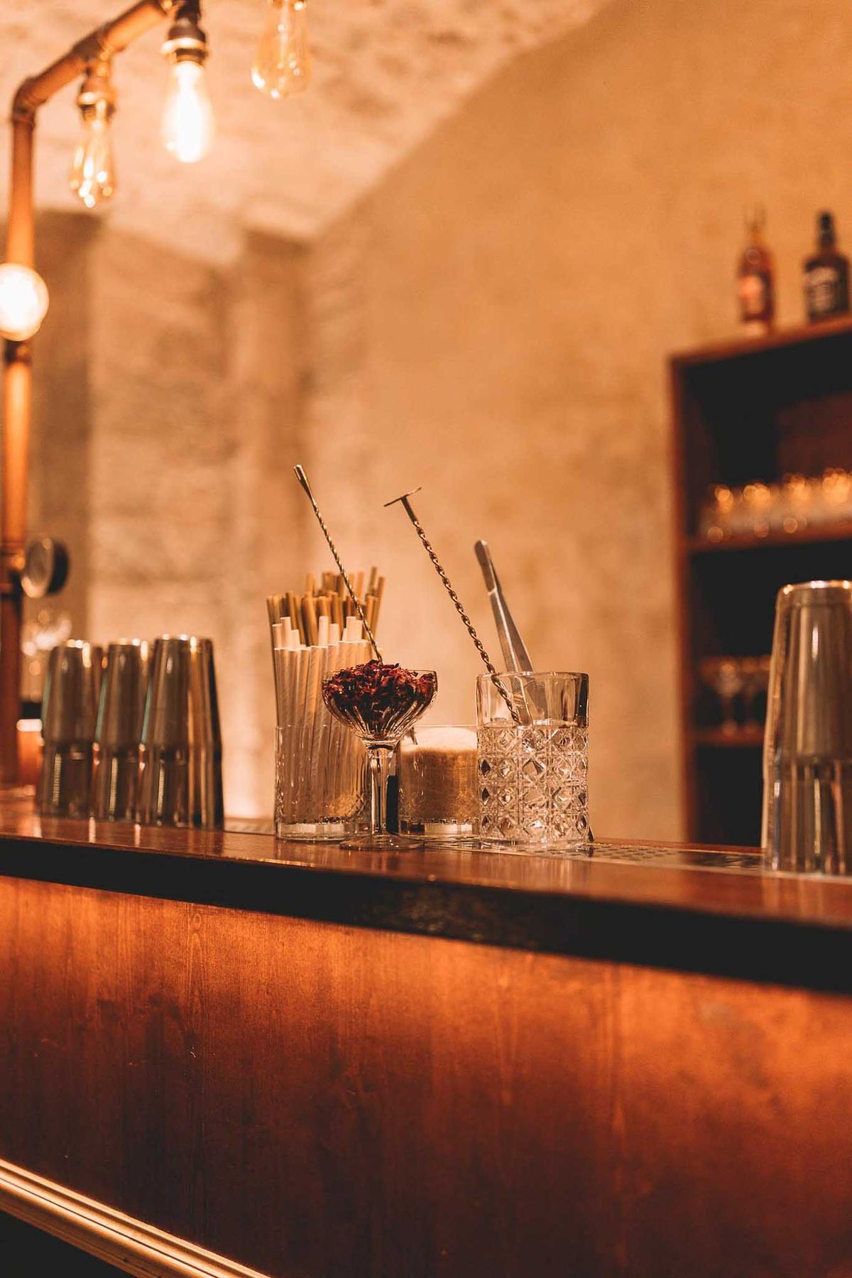 Close-up detail shot of a bar setup during what appears to be a wedding cocktail hour or reception, set in an indoor venue with exposed stone walls. The warm-toned wooden bar counter holds crystal cut-glass tumblers, metal cocktail shakers, bar spoons with twisted handles, paper straws in a tall glass, and a small coupe glass filled with dried dark red flower petals used as a garnish station. Overhead, bare Edison-style bulbs hang from copper pipe fixtures, casting warm amber light across the scene. In the background, a dark wood shelving unit displays spirit bottles including recognizable whisky labels, alongside glassware. The overall styling is industrial-rustic with a warm, candlelit palette of copper, amber, and gold tones.