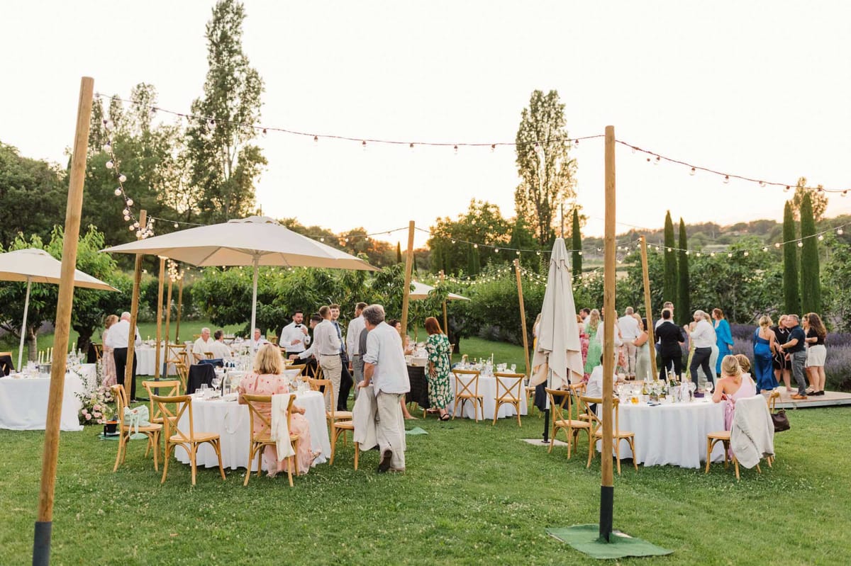 An outdoor wedding reception dinner is underway on a garden lawn at golden hour, with approximately 30–40 guests mingling and seated across multiple round tables. The tables are dressed in white linen and surrounded by natural wood cross-back chairs, with low floral centerpieces featuring blush pink and white blooms, candles, and glassware. Overhead, bistro globe string lights are strung between tall wooden poles, and large cream patio umbrellas provide shade over several tables. A small wooden dance floor is visible in the background on the right. The styling is relaxed and rustic-garden in tone, with a warm neutral palette of white, natural wood, and soft blush. Guests are dressed in a mix of smart-casual and formal attire, including floral print dresses, a blue dress, and a black tuxedo. The wide-angle shot captures the full reception layout from a slight elevation, conveying the overall scale and atmosphere of the setting.