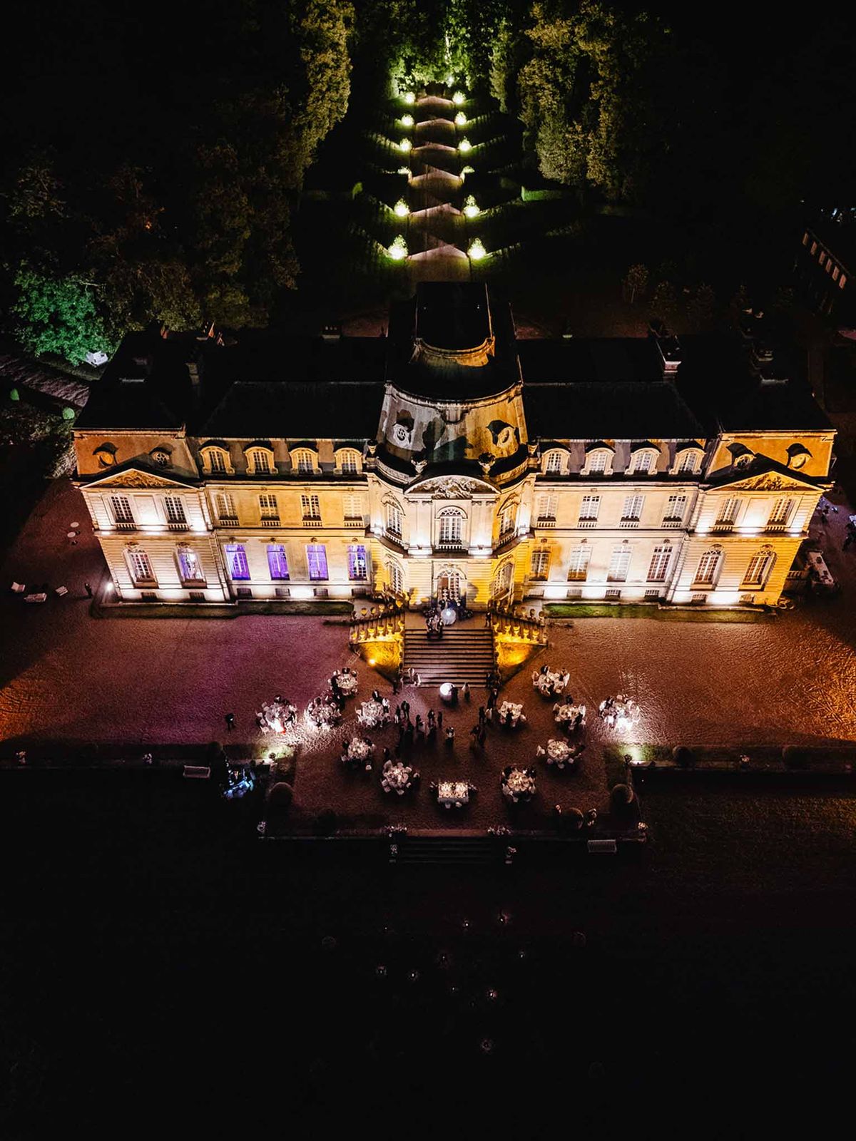 Aerial view of nighttime courtyard reception at chateau with 150-200 guests dining among candlelit tables and uplighting