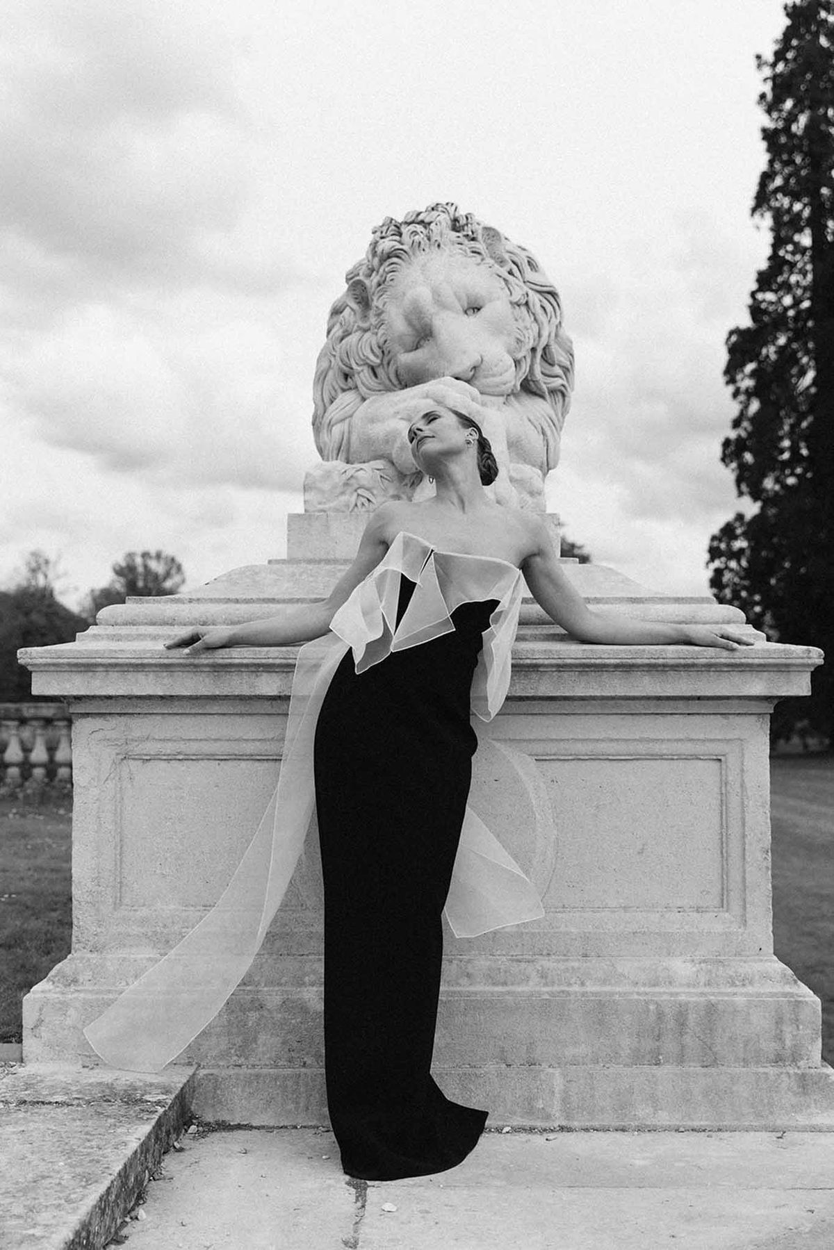 Bride in black strapless gown with white ruffled collar posing against stone lion sculpture in formal garden