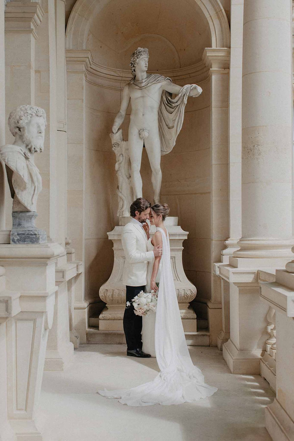 Bride in ivory silk gown and groom in cream jacket with burgundy tie in neoclassical gallery with marble busts
