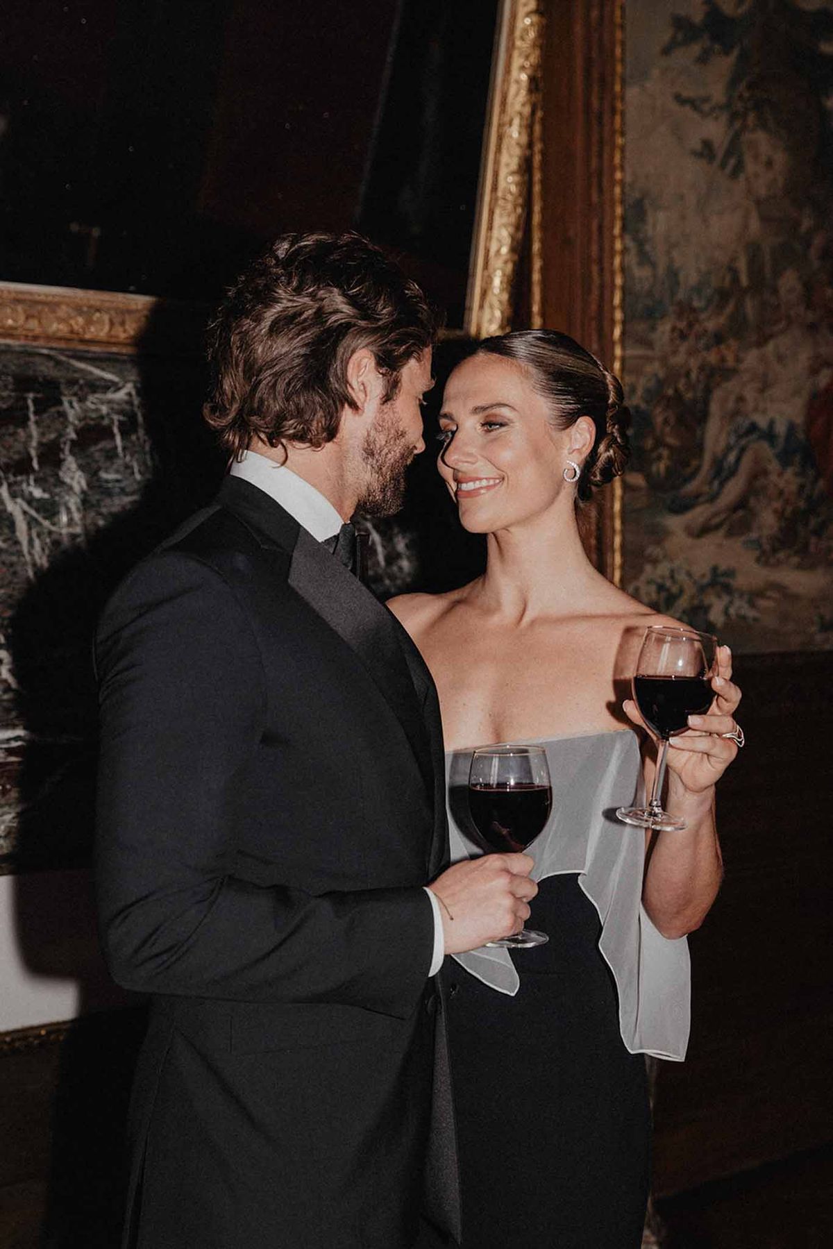 Bride in dove grey gown and groom in black tuxedo holding wine glasses during reception