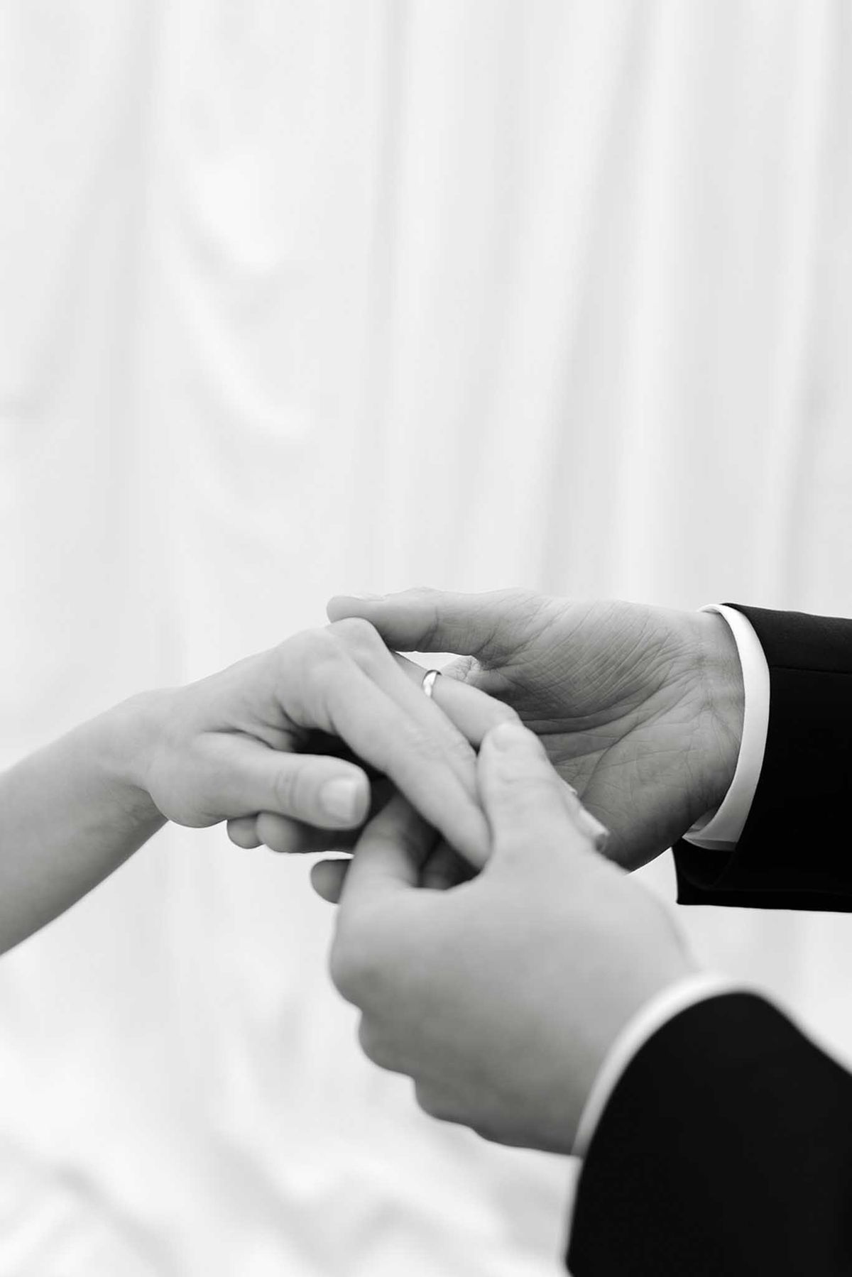 Close-up of bride and groom's hands during ring exchange, black and white, dark suit cuff against white dress