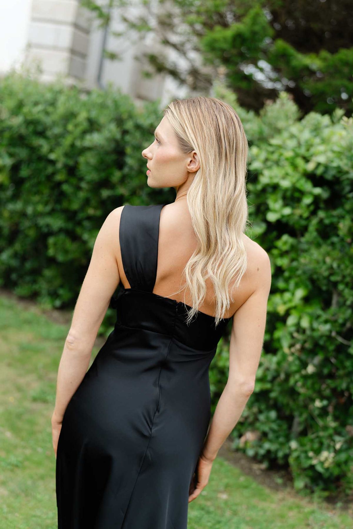 Female guest in black open-back dress photographed from behind in manicured garden with green hedges