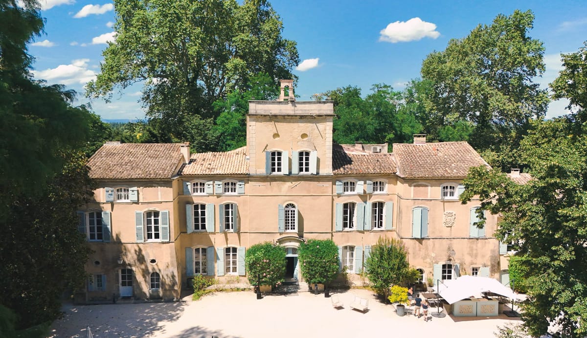 An aerial wide shot of a French château with warm terracotta-pink rendered façade, terracotta tile roof, and pale blue-grey shutters across multiple windows. The three-storey building features a central tower element with a sundial, arched entrance doorway framed by trimmed green hedging, and a stone coat-of-arms detail on the right wing. The gravelled forecourt in front of the building includes potted plants, a few white outdoor chairs, and a white market umbrella with two people visible nearby. The property is surrounded by mature deciduous trees on all sides. Potential venue feature image.