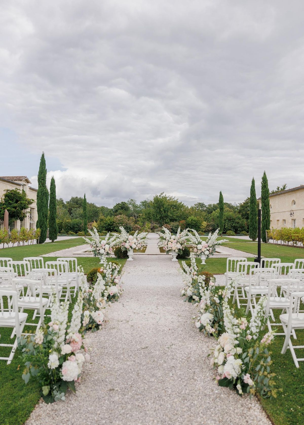 An outdoor wedding ceremony setup photographed from the back of the aisle looking toward the altar, captured as a wide shot. The ceremony is set on a gravel aisle flanked by rows of white Chiavari chairs arranged on a formal garden lawn, with the buildings of what appears to be a French chateau or domaine visible on either side. The aisle is lined with low floral arrangements featuring white peonies, blush pink roses, white delphiniums, and eucalyptus greenery placed directly on the ground at regular intervals. At the far end, large white pedestal urns hold full, overflowing floral arrangements in the same white and blush palette, framing a staircase altar area. The overall decor palette is white and soft blush pink with green foliage, styled in a classic formal manner. Potential venue feature image.
