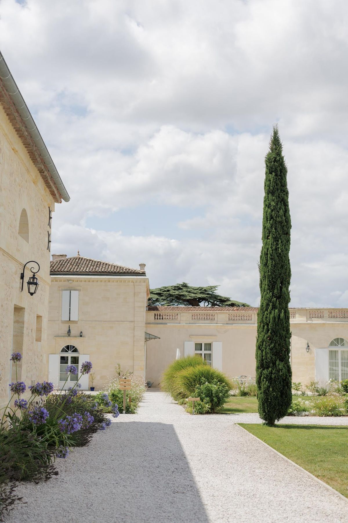 Cream limestone chateau courtyard with terracotta roofs, white shutters, cypress, and purple agapanthus