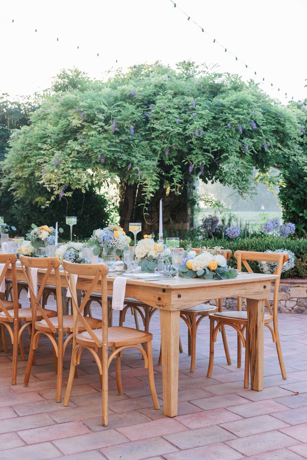 Reception tables under wisteria-covered pergola with peach roses, blue hydrangeas, and eucalyptus centerpieces on farmhouse tables