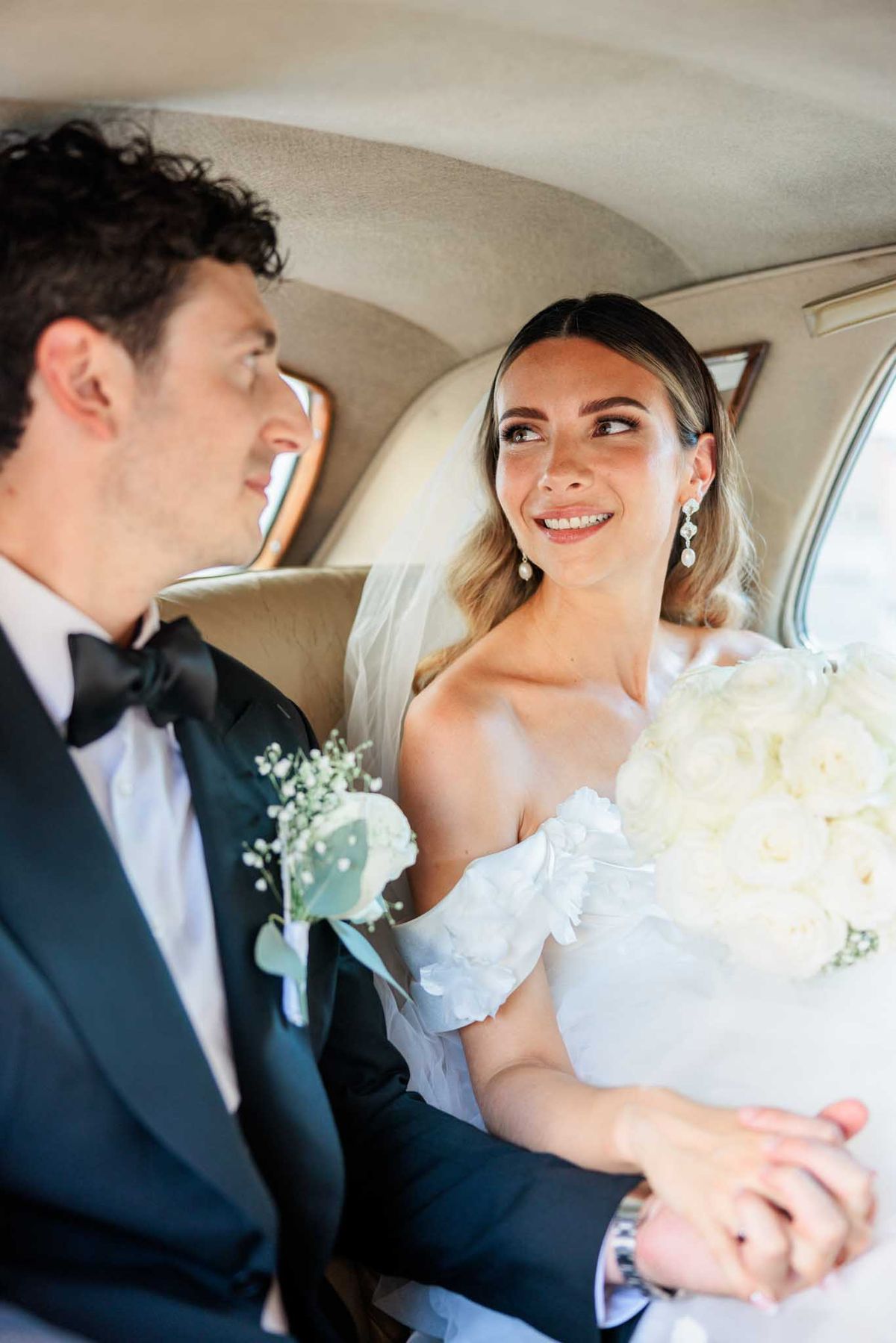 Bride in off-shoulder ruffled gown and groom in tuxedo sharing an intimate moment in back seat of wedding car
