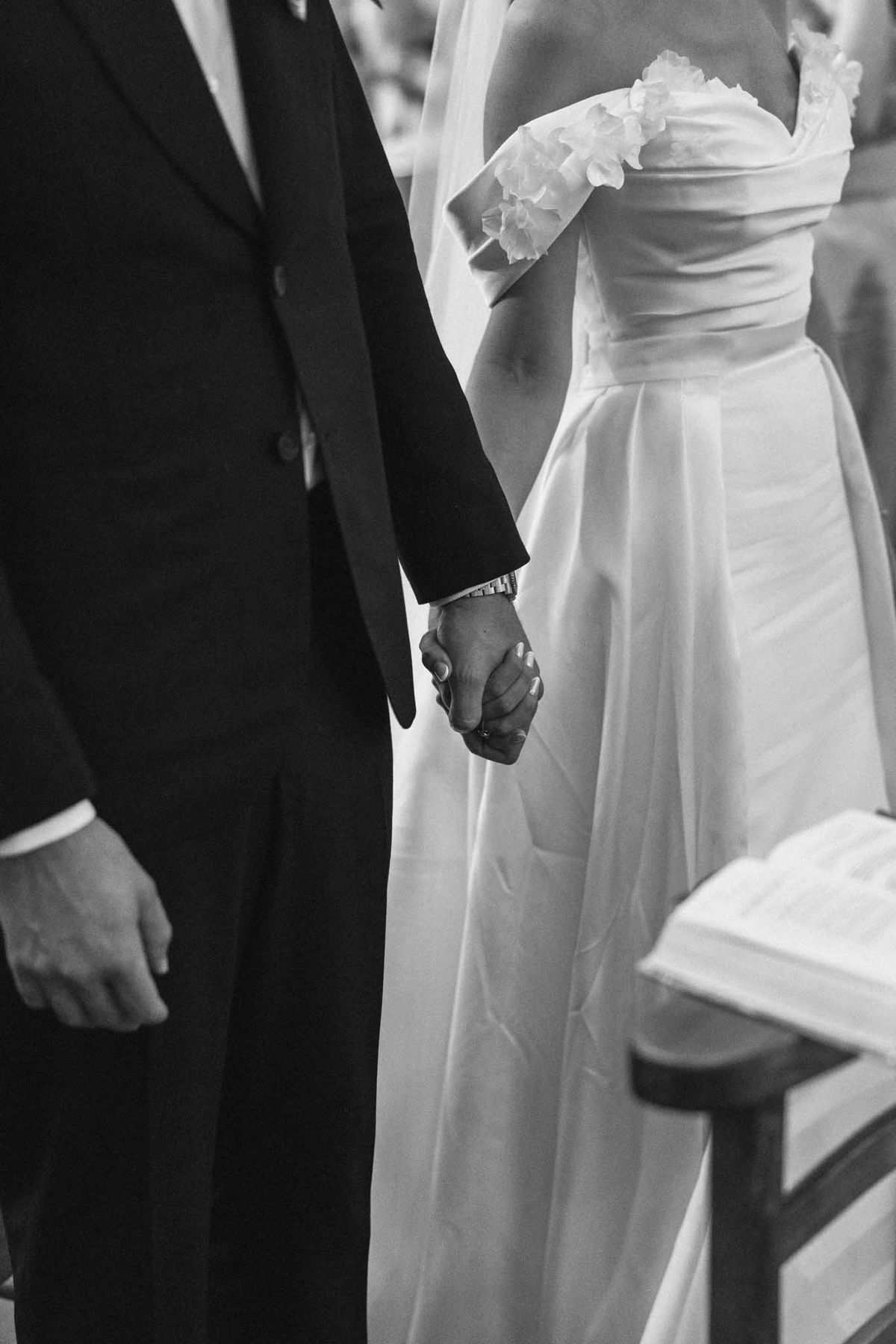Black and white close-up of bride and groom holding hands during ceremony, groom's watch visible, officiant behind